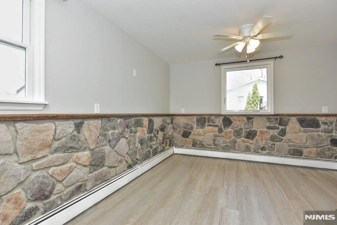 Empty room with stone wainscoting, light wood flooring, and white walls, featuring a ceiling fan and two windows.