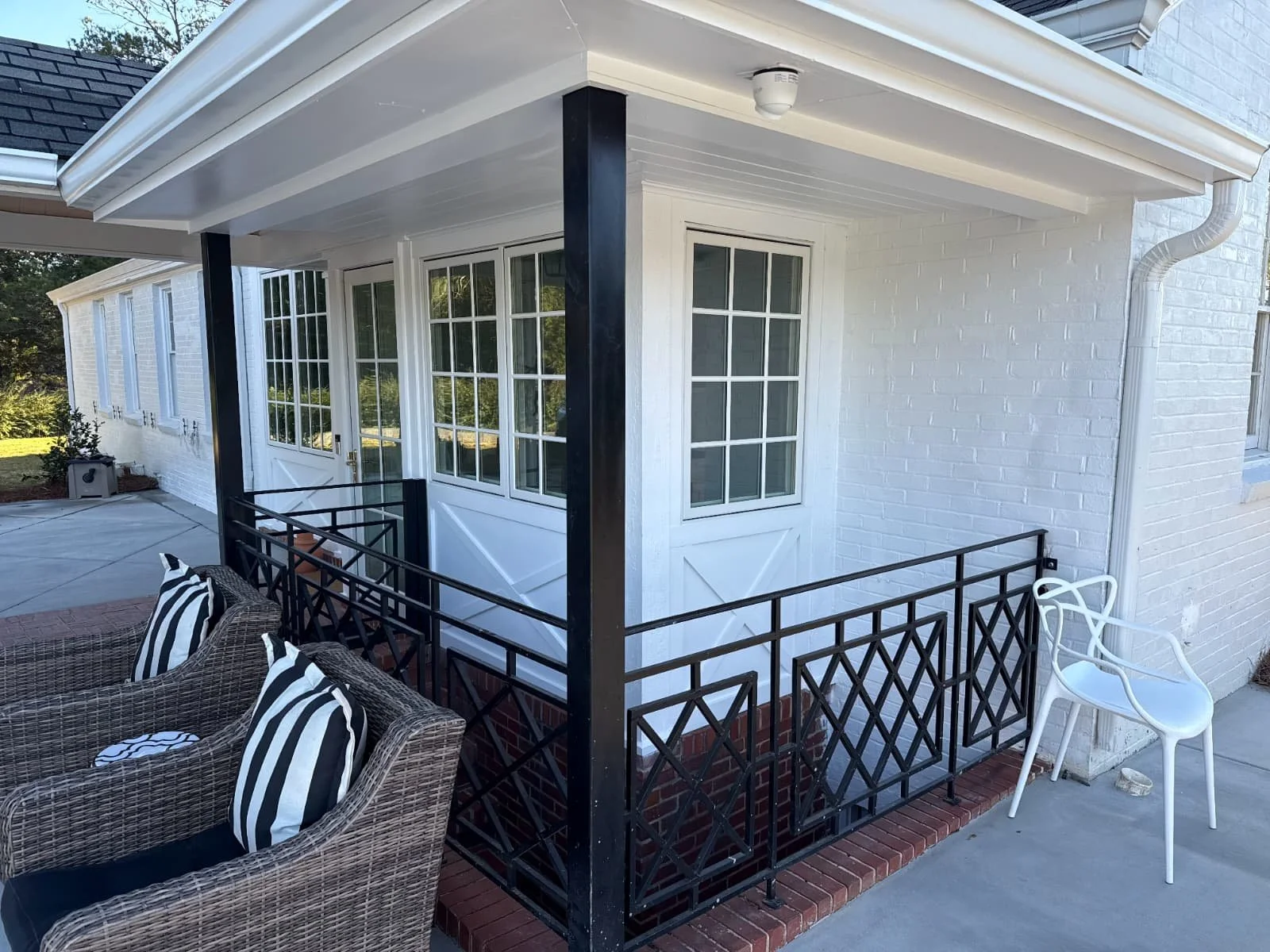 A small porch enclosed with black wrought iron railing, attached to a white brick house with multiple large windows. There is outdoor furniture, including a wicker chair with black and white striped cushions, and a white plastic chair. The porch has 