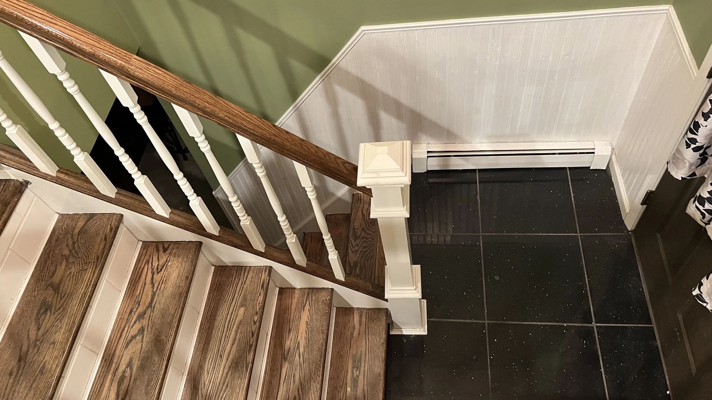 Wooden stairs with a brown handrail and white balusters, black tiled floor, green and white walls with beadboard paneling, and a floral curtain.