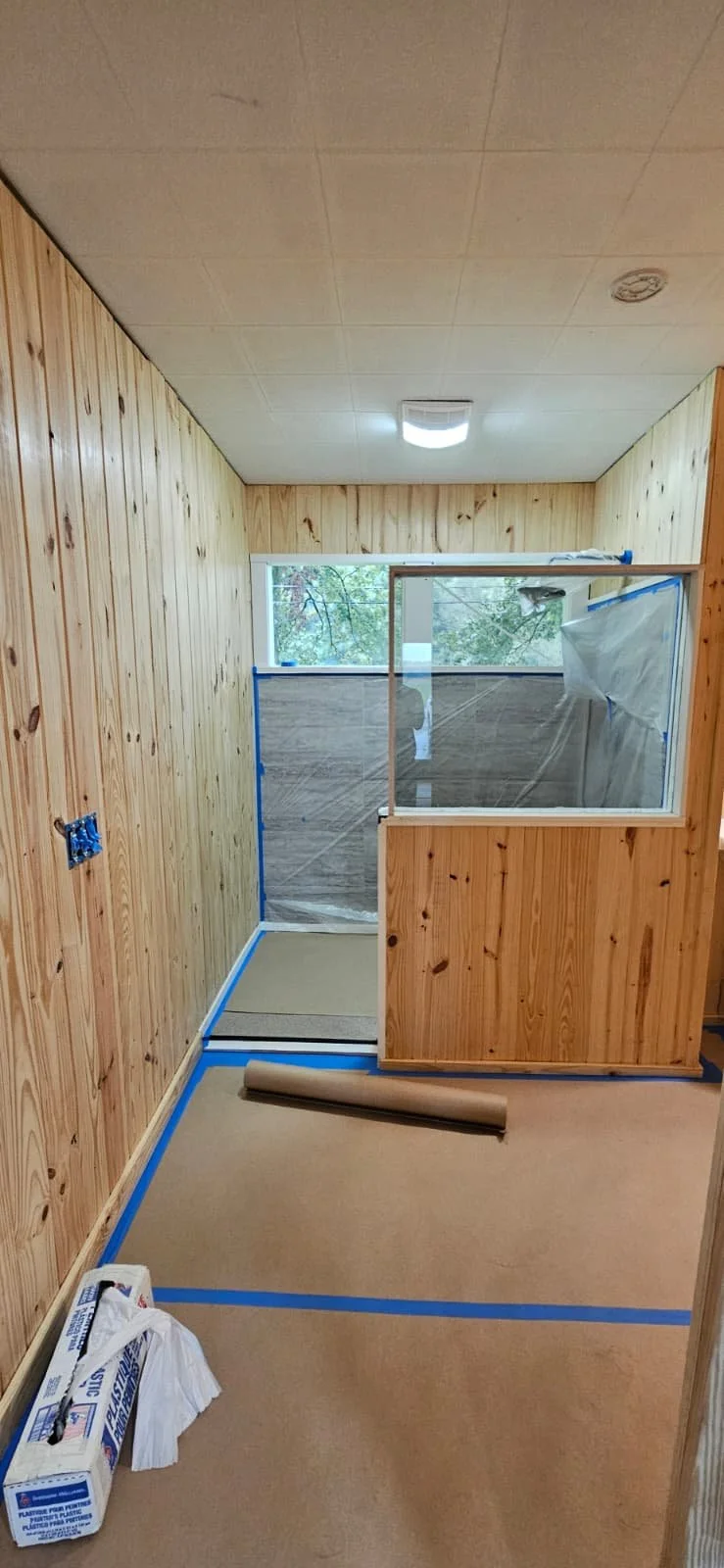 A small room under construction with wood-paneled walls, beige carpeted floor, and a window at the back showing trees outside. A partially assembled glass shower enclosure is visible, with tools and plastic sheeting. Blue painter's tape outlines the 