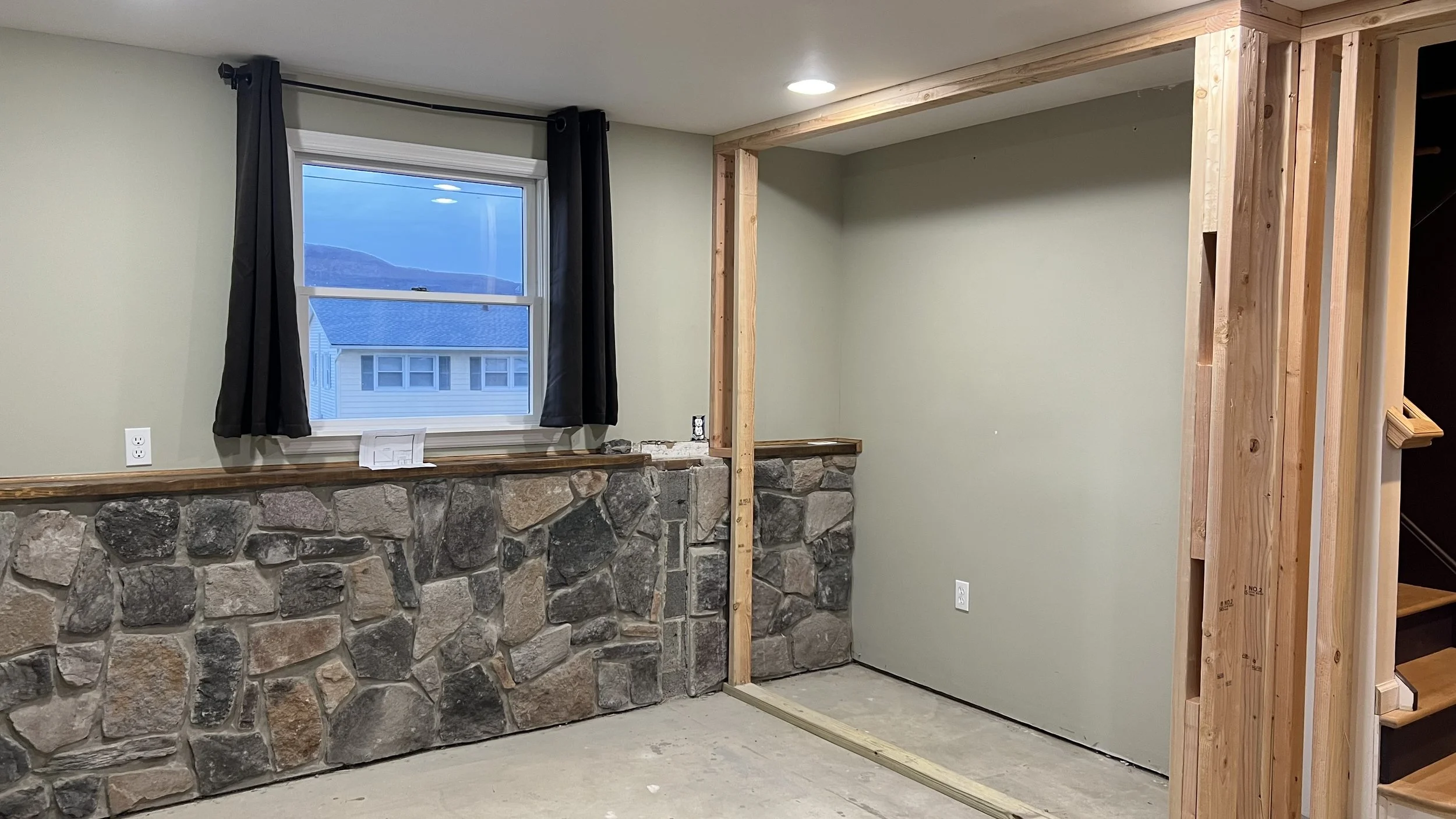Interior of a house under construction with a stone wall below a window, black curtains, and exposed wooden framing next to a closed wall.
