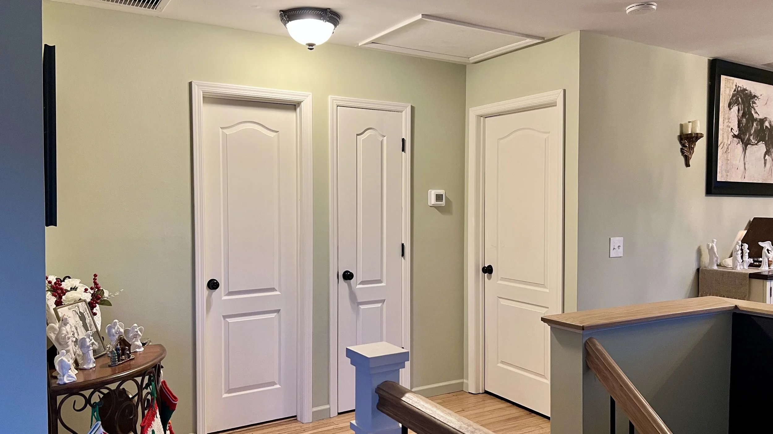 Interior of a house showing three white interior doors with black doorknobs, a wall-mounted thermostat, and a framed painting of a black horse. A small table with holiday decorations and figurines is visible in the corner.