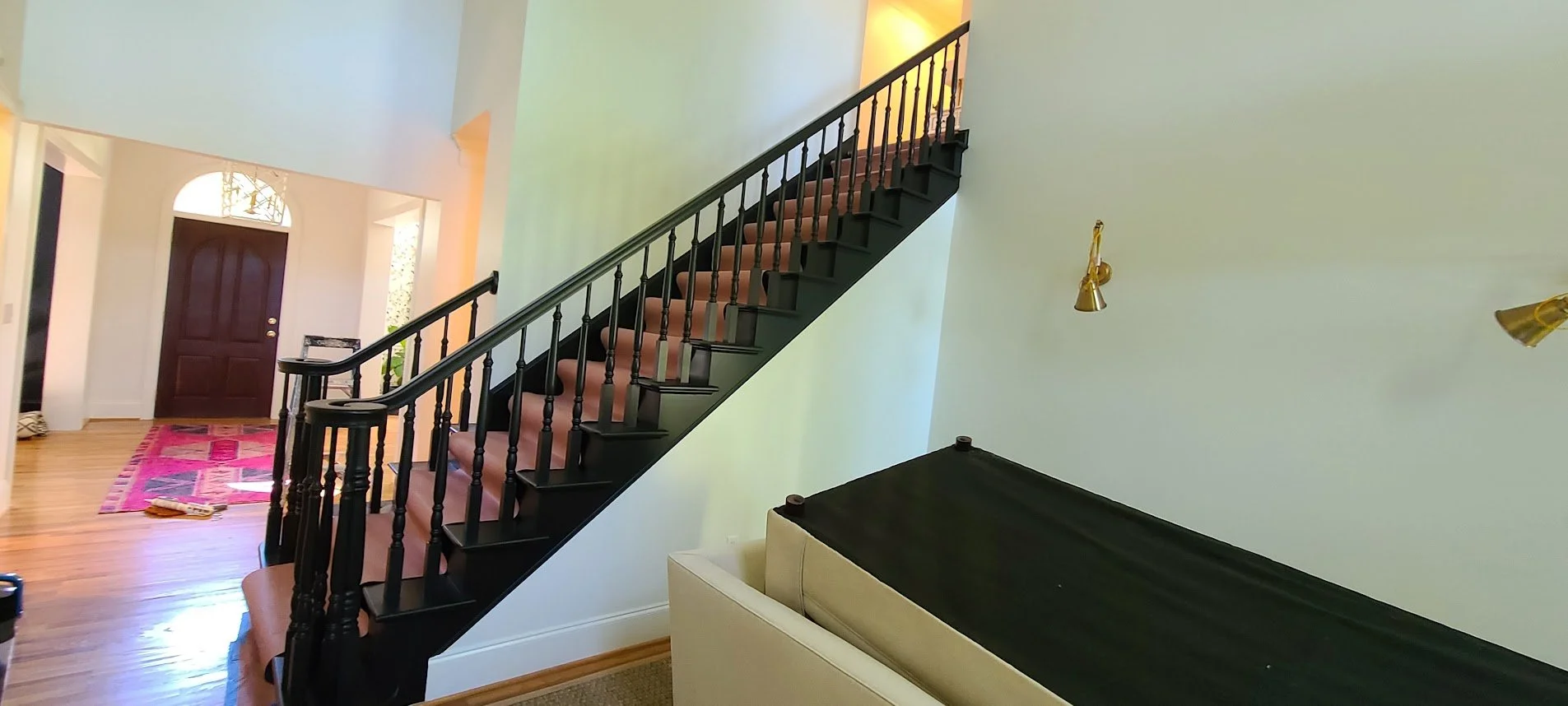 Interior view of a house showing staircase with black banister, beige carpeted steps, wooden floor entrance area with a red and pink patterned rug, front door with an arched window, and a light-colored wall with gold wall lamps.