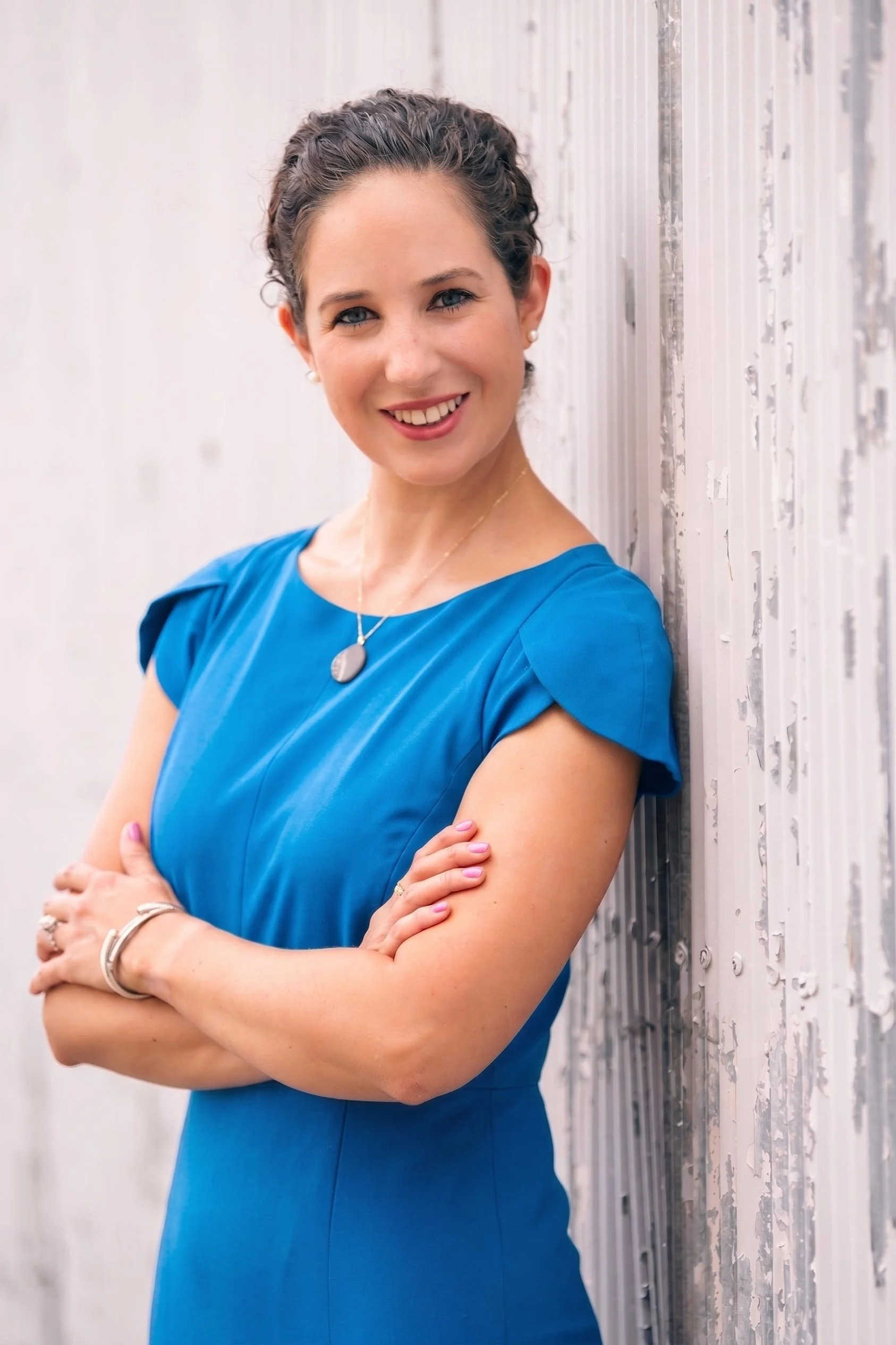 A woman with blue eyes and curly dark hair smiling and leaning against a white, textured wall with her arms crossed, wearing a blue dress and jewelry.