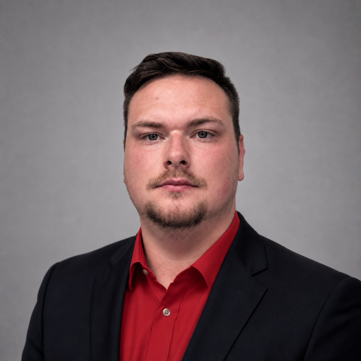 Professional headshot of a man with dark hair, a goatee, and a mustache, wearing a black suit jacket and a red dress shirt, against a plain gray background.