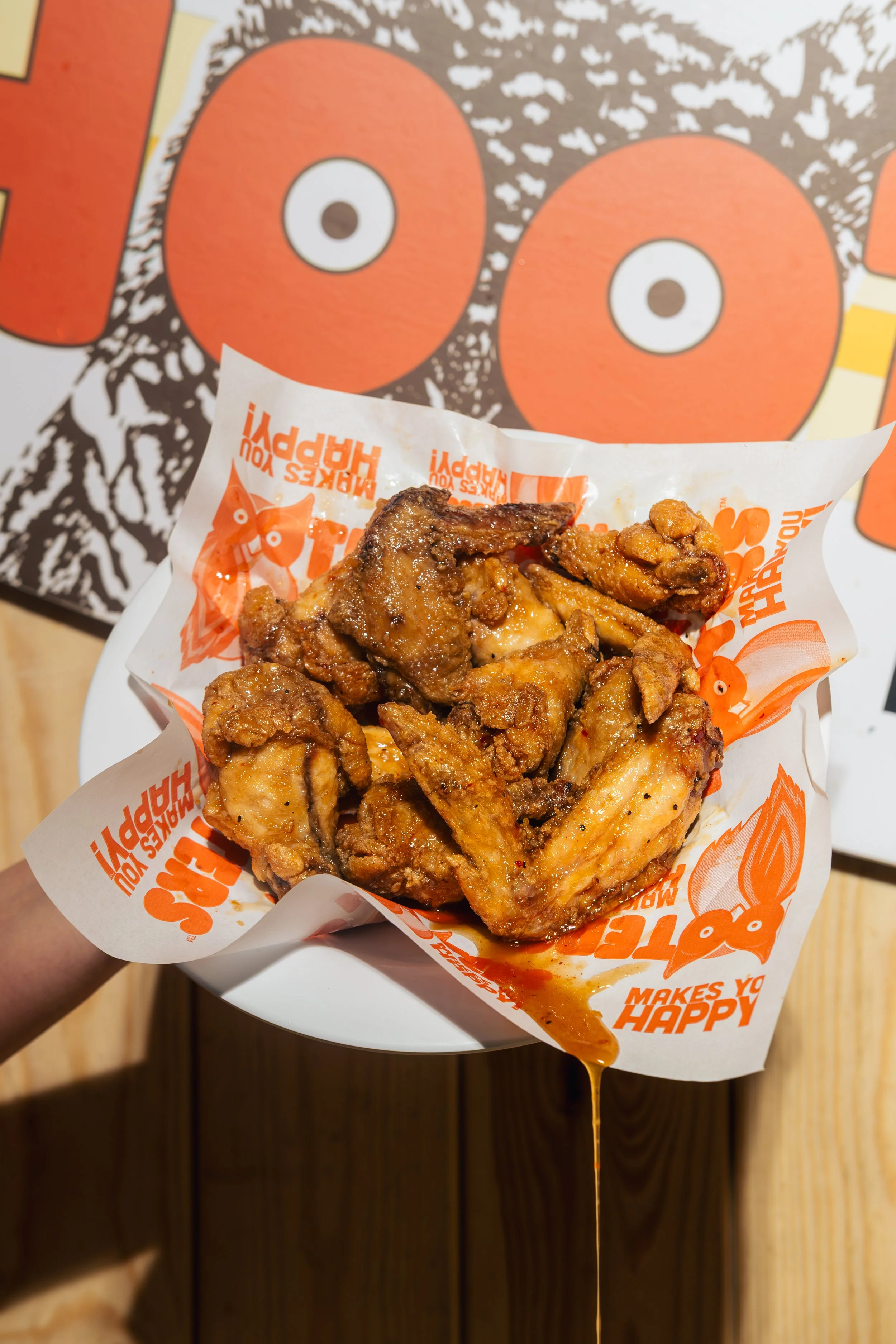 A serving of fried chicken pieces in a paper wrapper with bright orange and white text, held above a white plate, with a cartoon poster of a face with large orange cheeks and wide eyes in the background.