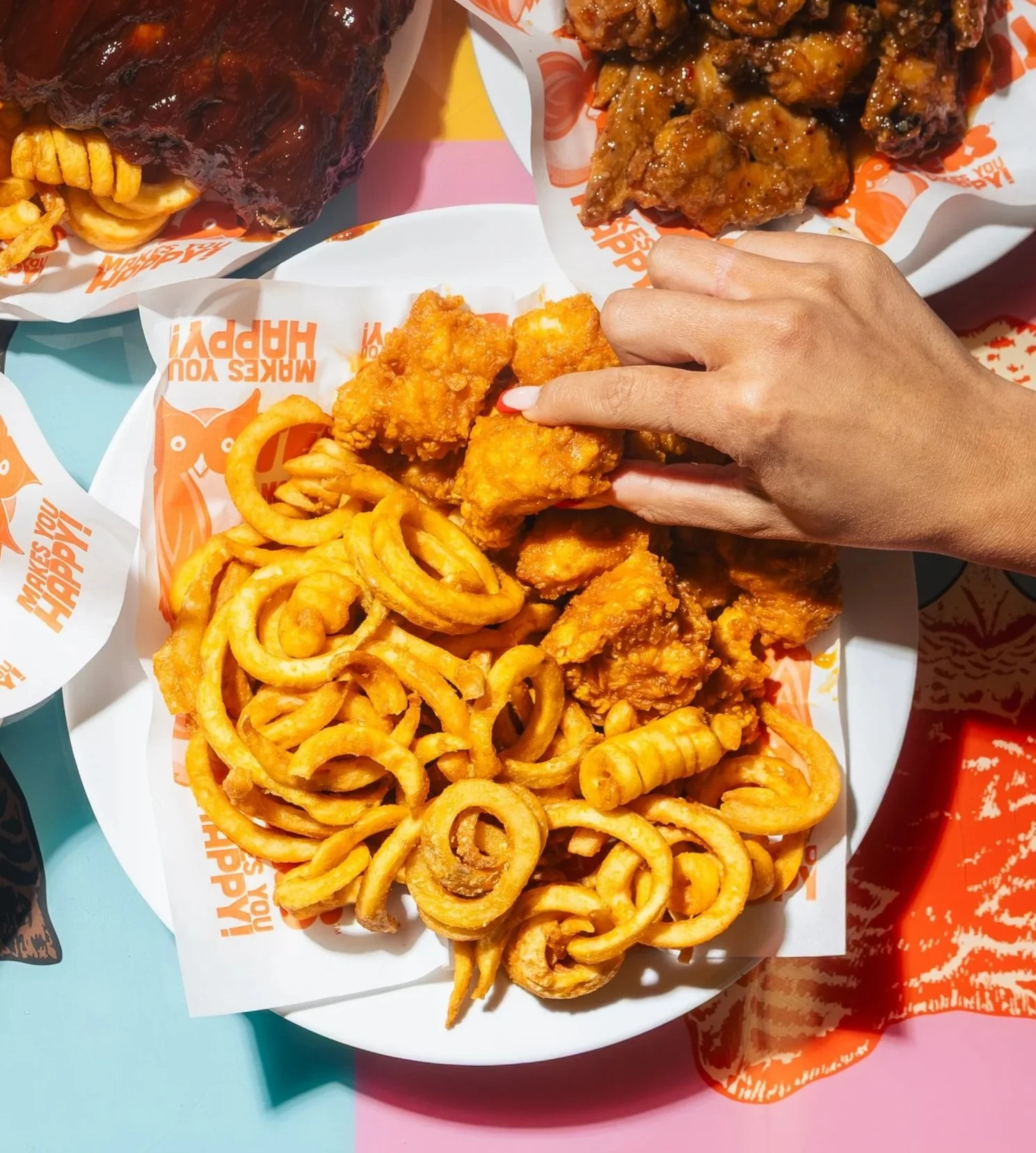 A plate of fried onion rings in the foreground, with spicy crispy chicken pieces, and barbecue ribs in paper trays in the background, on a colorful table.