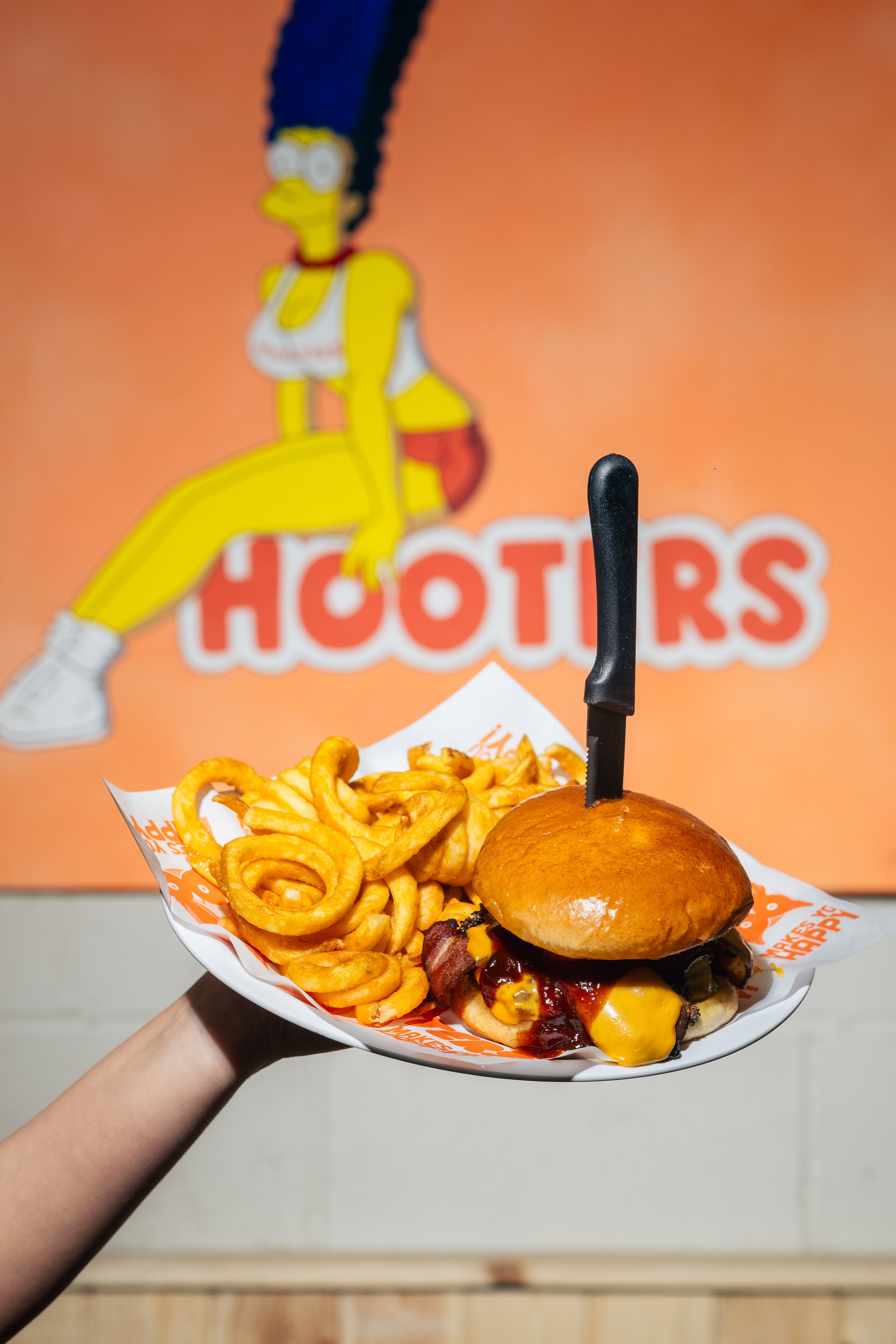 A person holding a plate of fast food consisting of curly fries and a cheeseburger with bacon and cheese, in front of a bright orange 'Hooters' sign with a cartoon waitress logo.