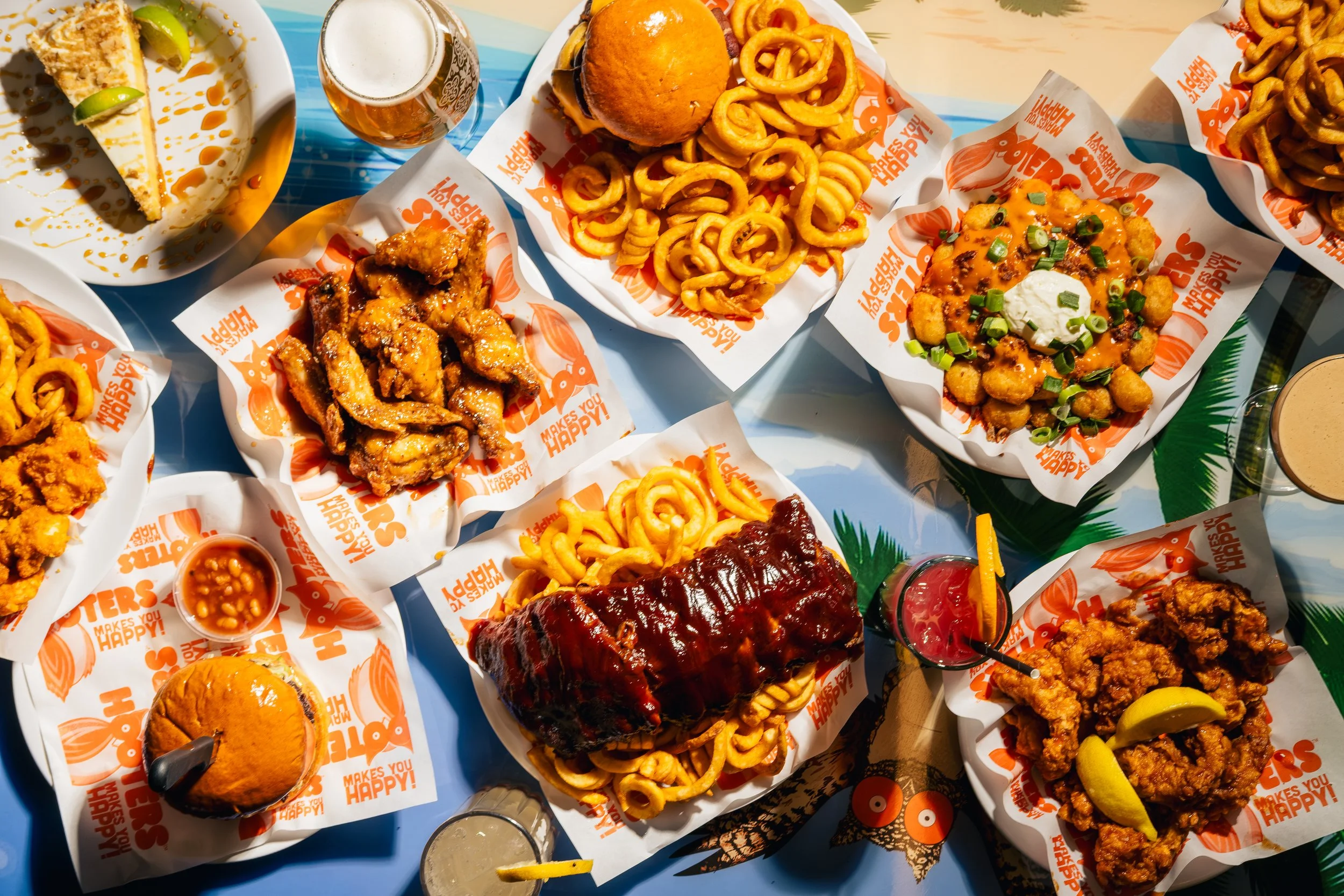 A spread of American-style fast food including fried chicken, curly fries, cheeseburgers, a slice of cheesecake, and various dipping sauces, on a colorful table.
