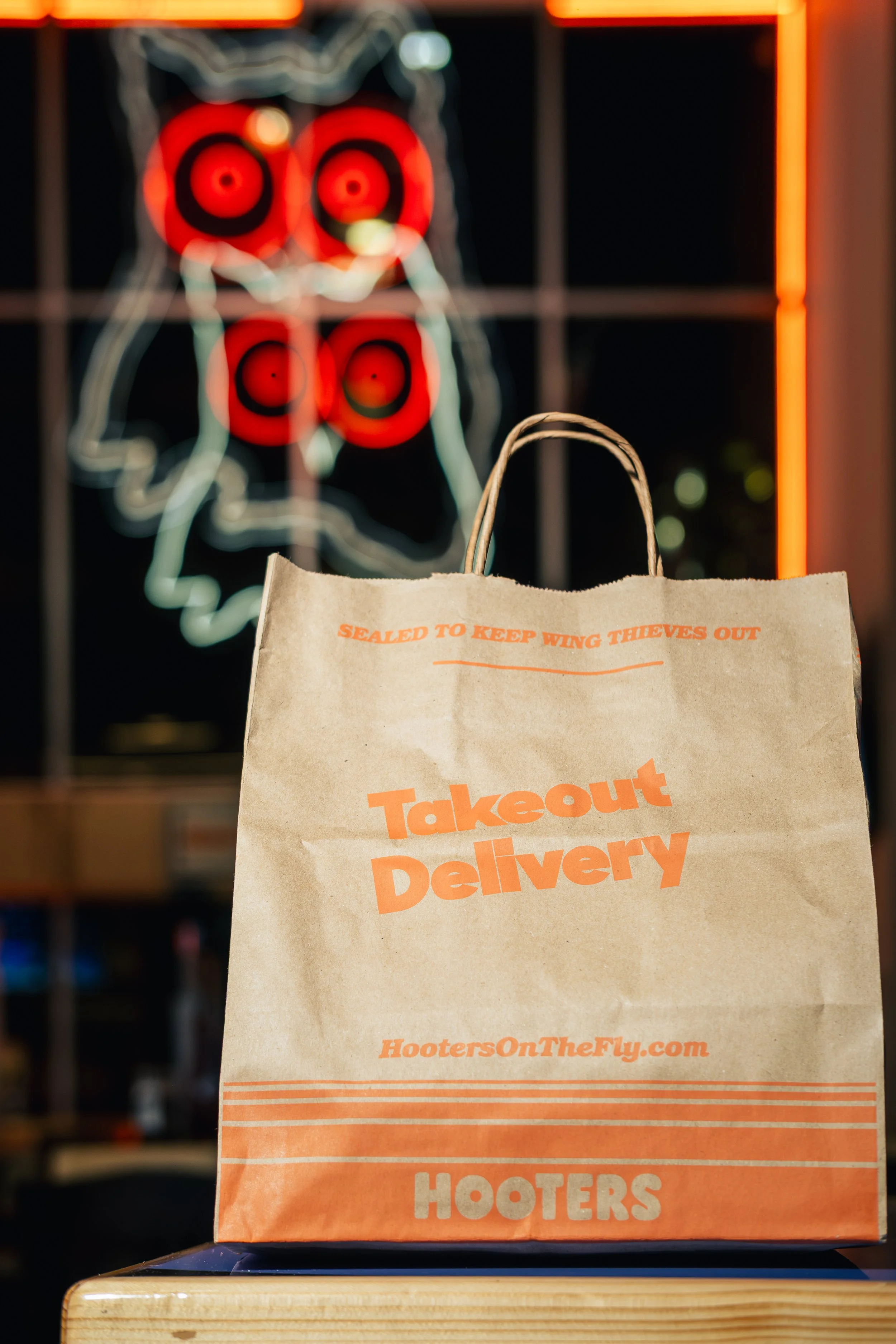 A brown paper takeout bag from Hooters on the Fly sits on a table, with a neon owl sign in red and white lights in the background.