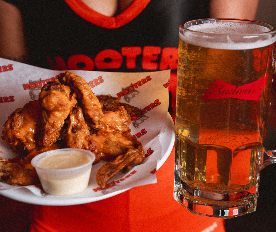 A person holding a tray with fried chicken wings and a cup of dipping sauce, next to a large mug of beer at Hooters restaurant.