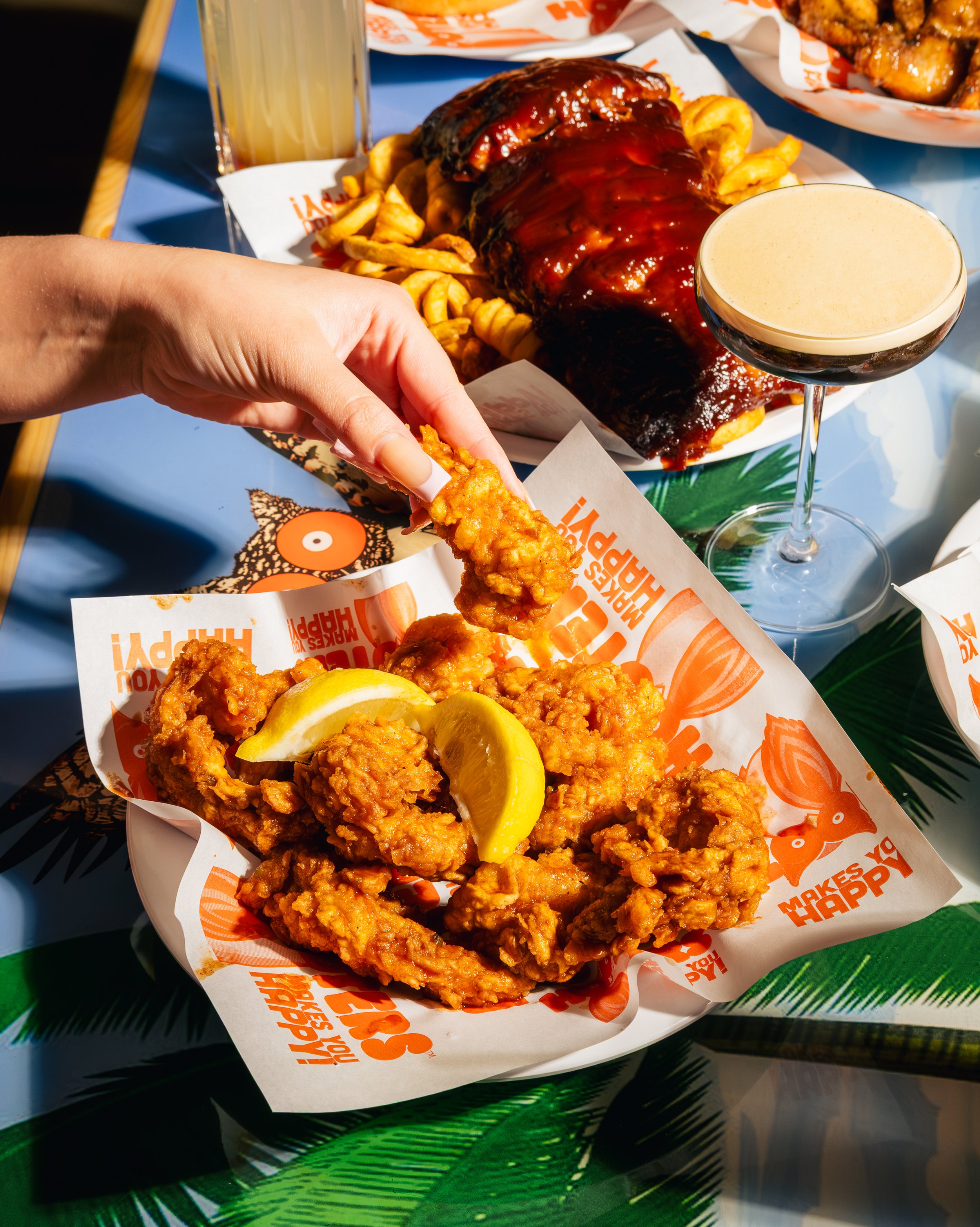 Fried chicken tenders with lemon wedges, a burger with barbecue sauce and curly fries, a glass of beer, and a foam-topped cocktail on a tropical-themed table.