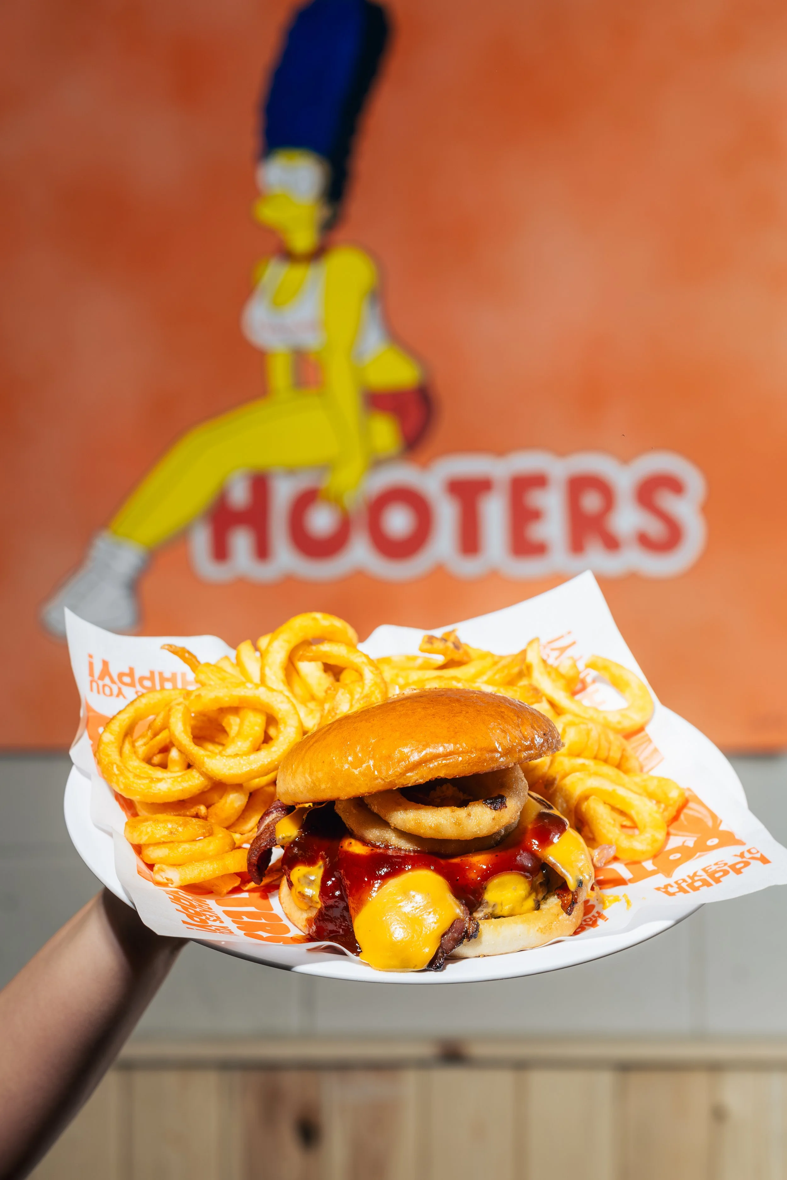 A plate of fast food with a cheeseburger topped with bacon, onions, sauce, and cheese, served with curly fries, held in front of a Hooters restaurant background.