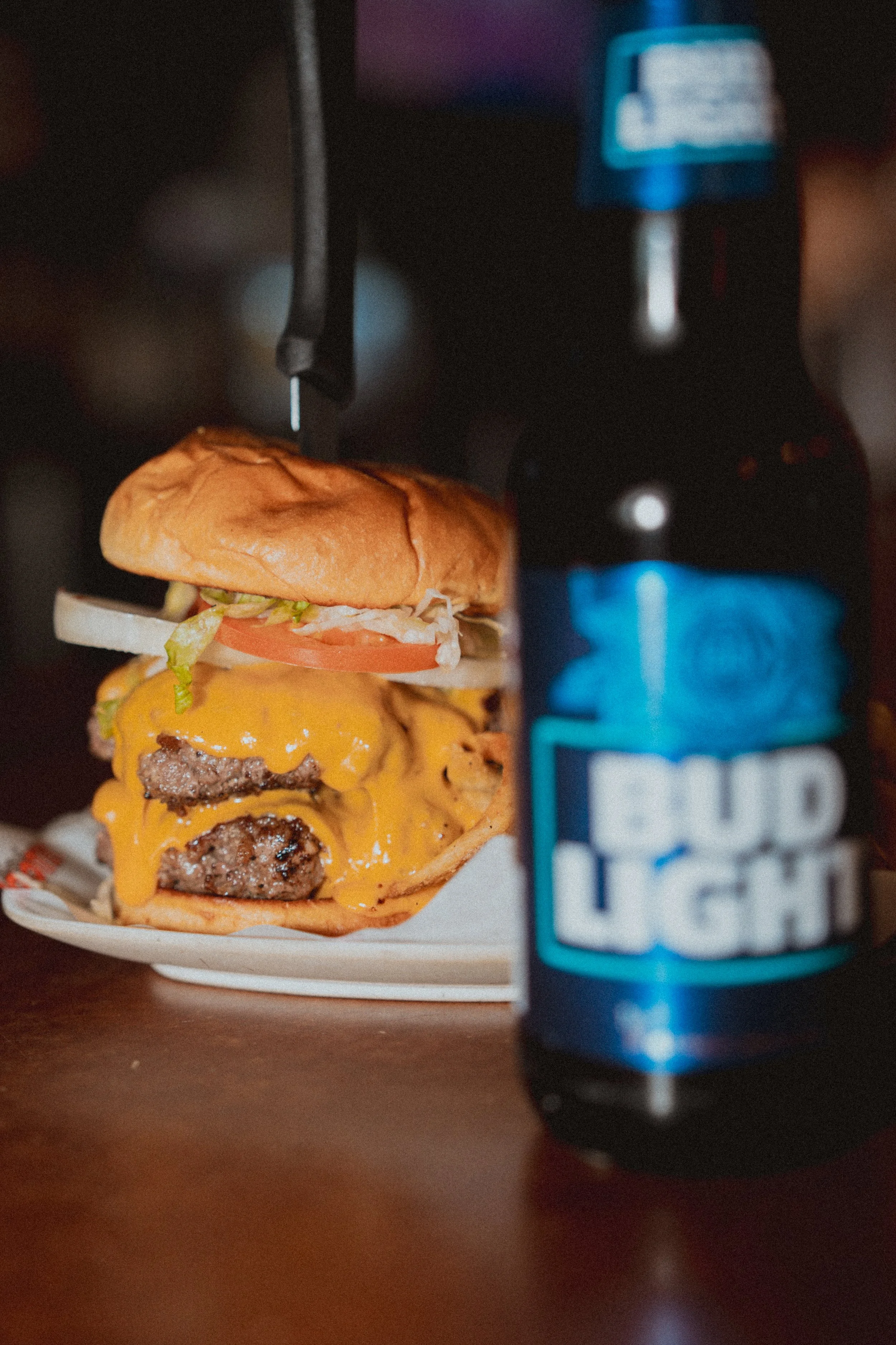 Close-up of a cheeseburger with lettuce, tomato, onion, and melted cheddar cheese, placed on a plate, with a blurry blue and black soda bottle in the foreground.