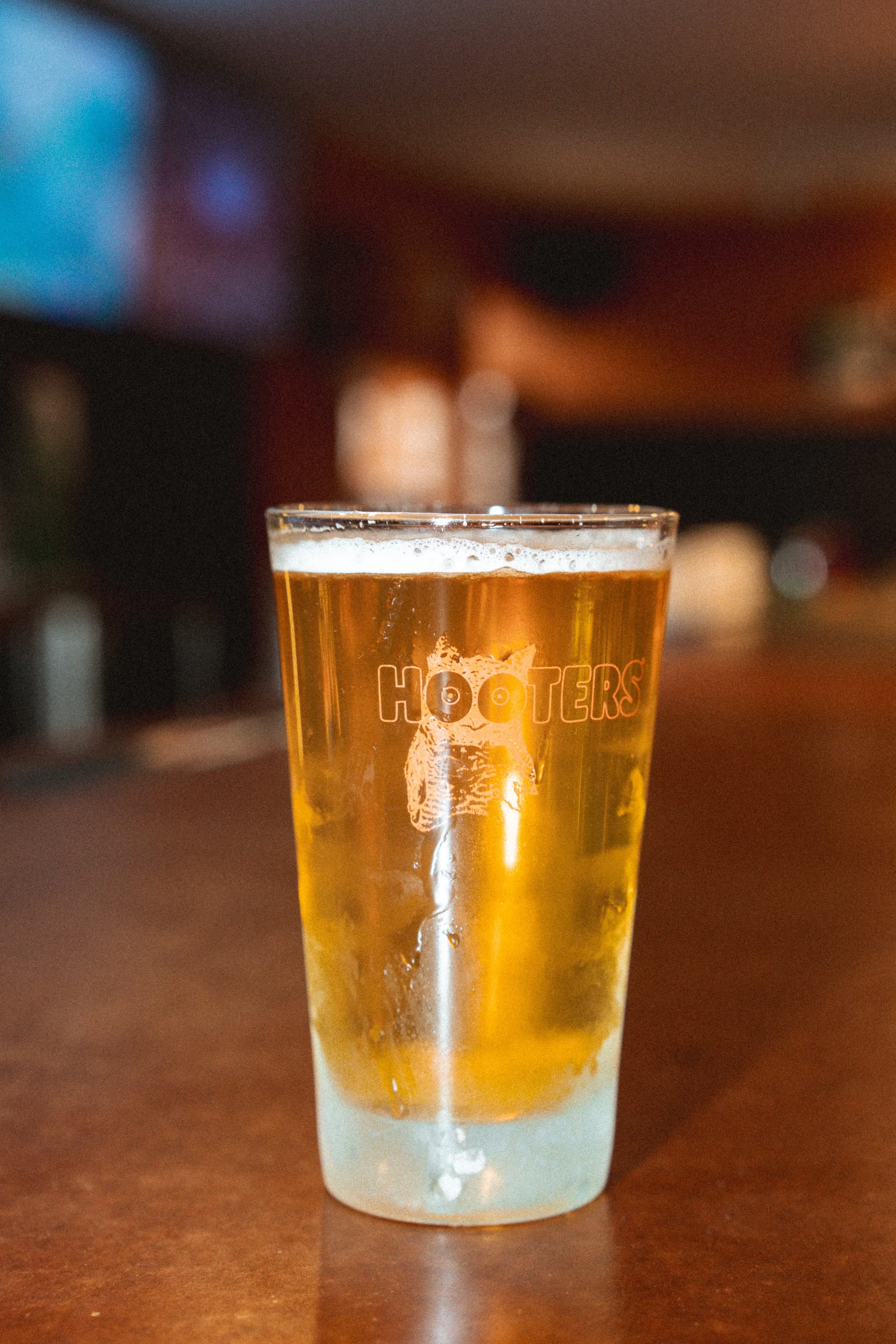 A glass of beer with the HOOTERS logo on it, sitting on a wooden surface in a bar or restaurant setting.