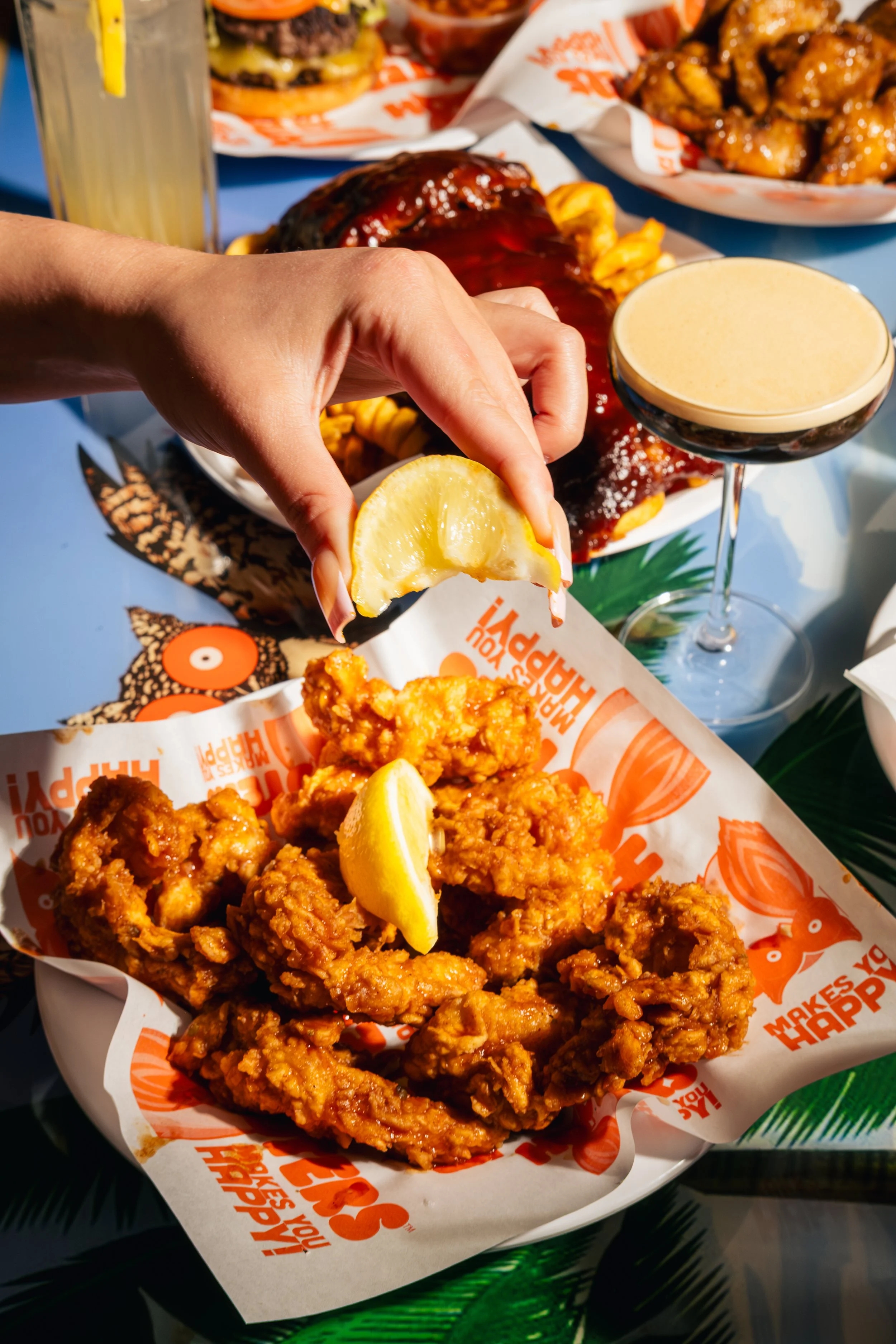 Fried chicken with lemon wedges, dipping sauce, and beer on a table with fast food items in the background.