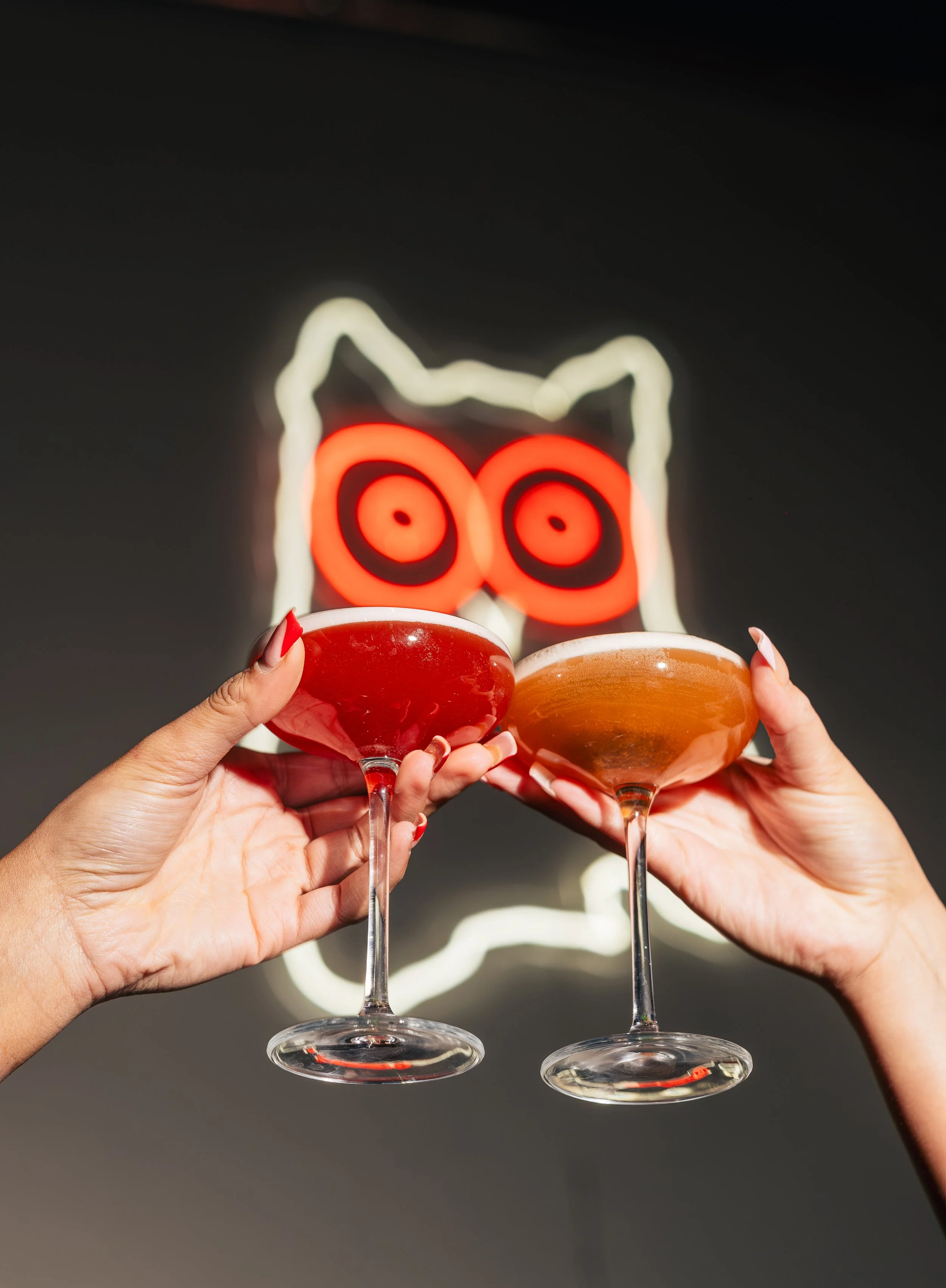 Two people holding cocktail glasses with red and orange drinks, with a neon sign of an owl in the background.