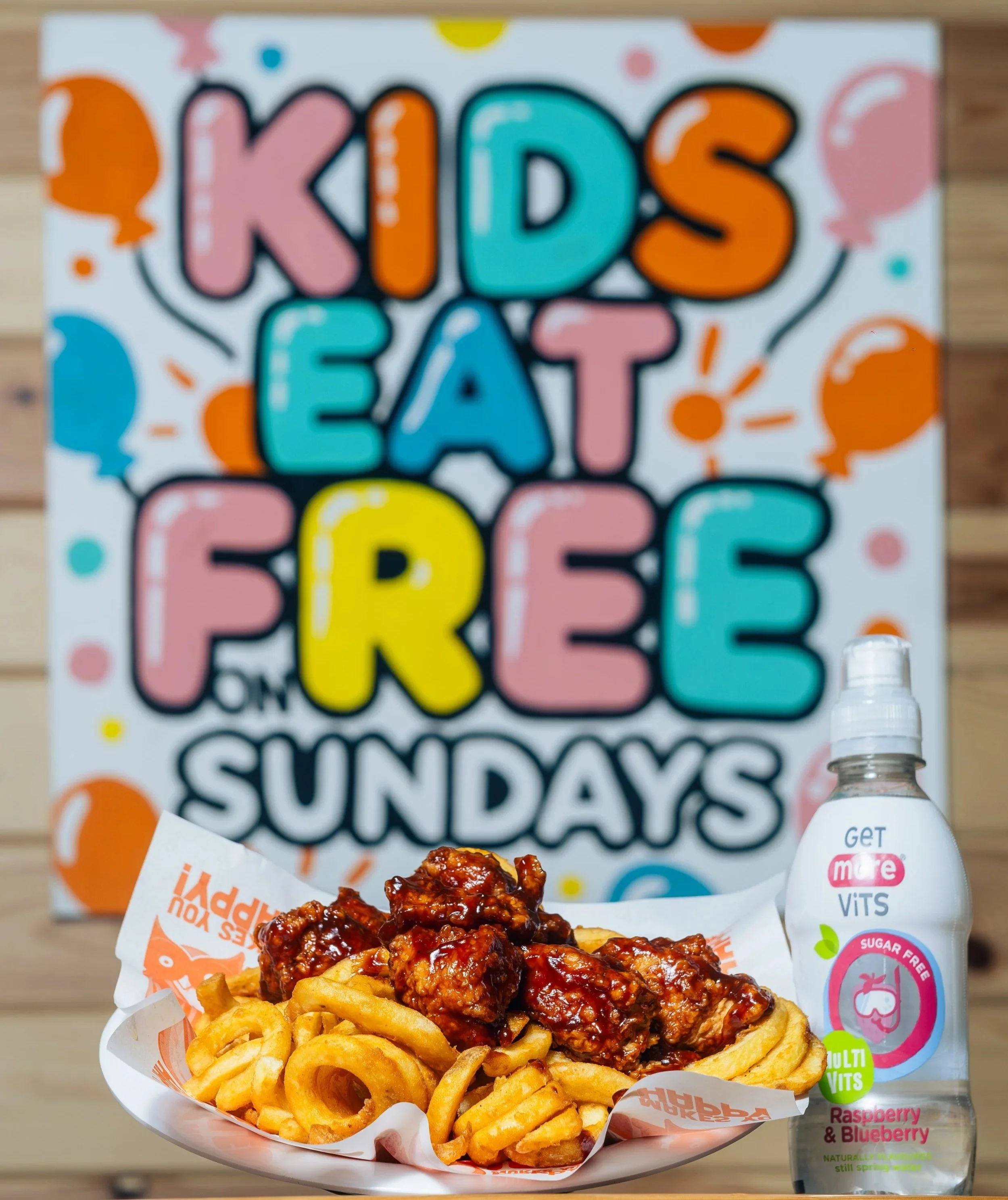 A plate of curly fries topped with glazed chicken pieces in sauce, set in front of a blurred background containing a colorful poster and a water bottle.