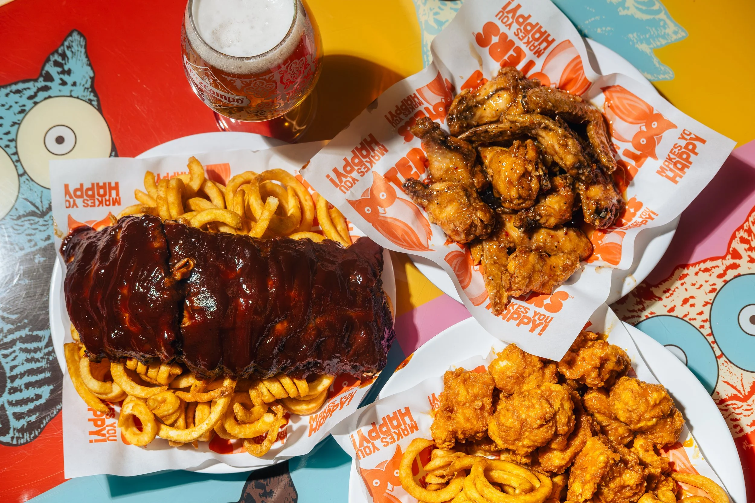 Fried chicken, curly fries with barbecue sauce, and fried chicken with hot sauce on trays at a colorful restaurant table.