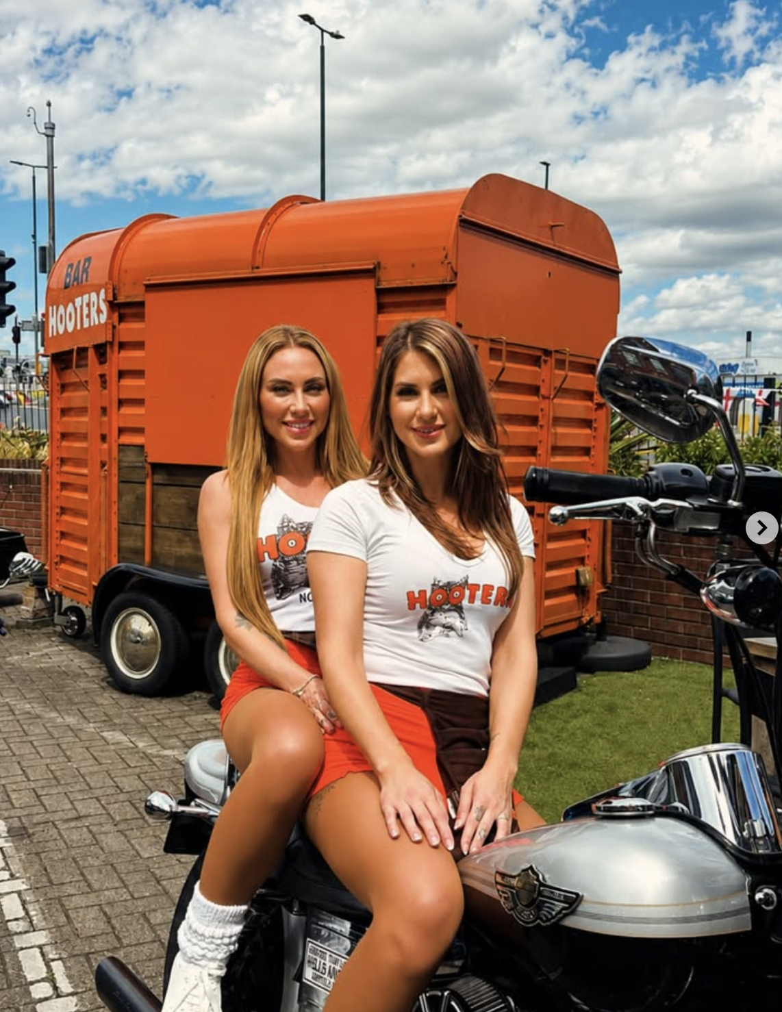 Two women sitting on a motorcycle, both wearing white T-shirts with Hooters logo, outdoors in front of an orange food trailer, under a partly cloudy sky.