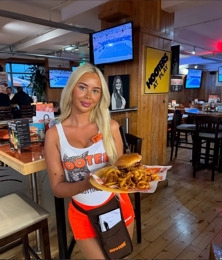 A Hooters waitress with long blonde hair holding a tray of curly fried onion rings and a burger inside a restaurant with wooden walls, televisions, and multiple tables.