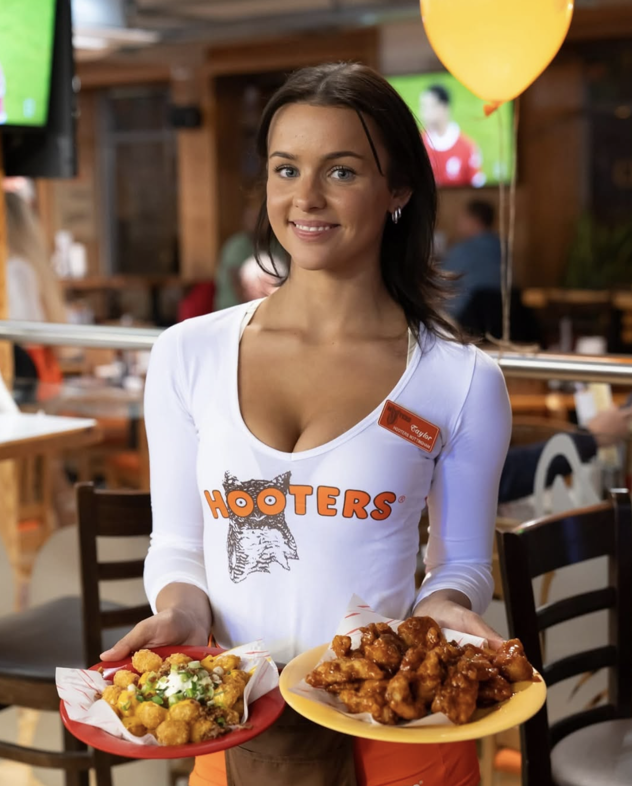 A young woman wearing a Hooters uniform serving plates of chicken wings and fried food at a restaurant.