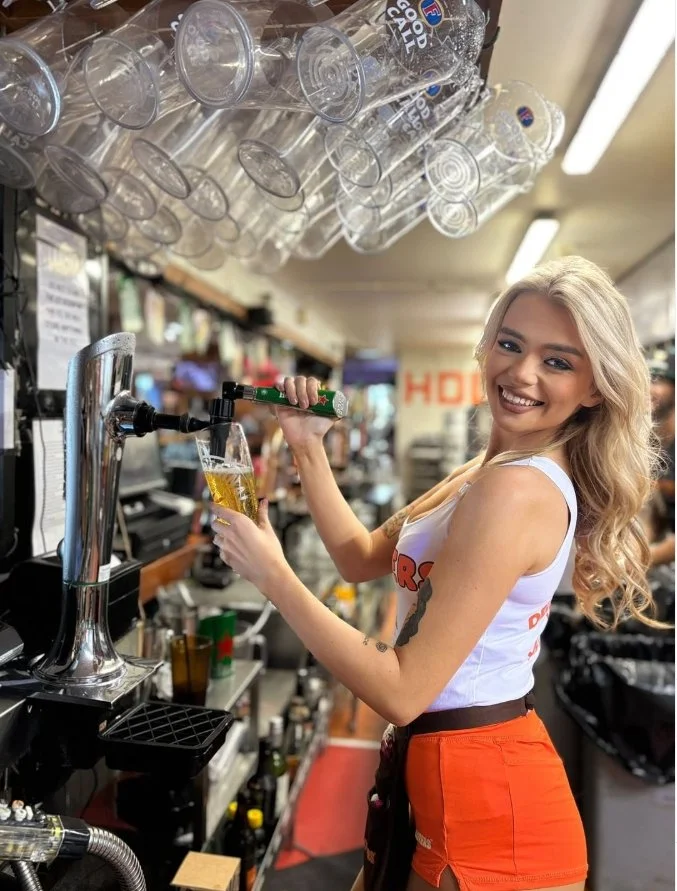 A smiling blonde woman with wavy hair pours a draft beer into a glass at a bar, with empty glasses hanging above her.