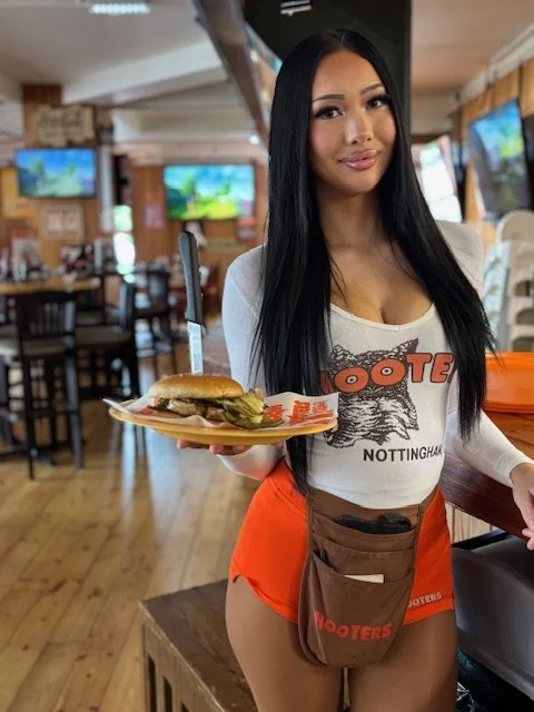 A young woman in a Hooters uniform holding a plate with a burger and knife inside a restaurant.