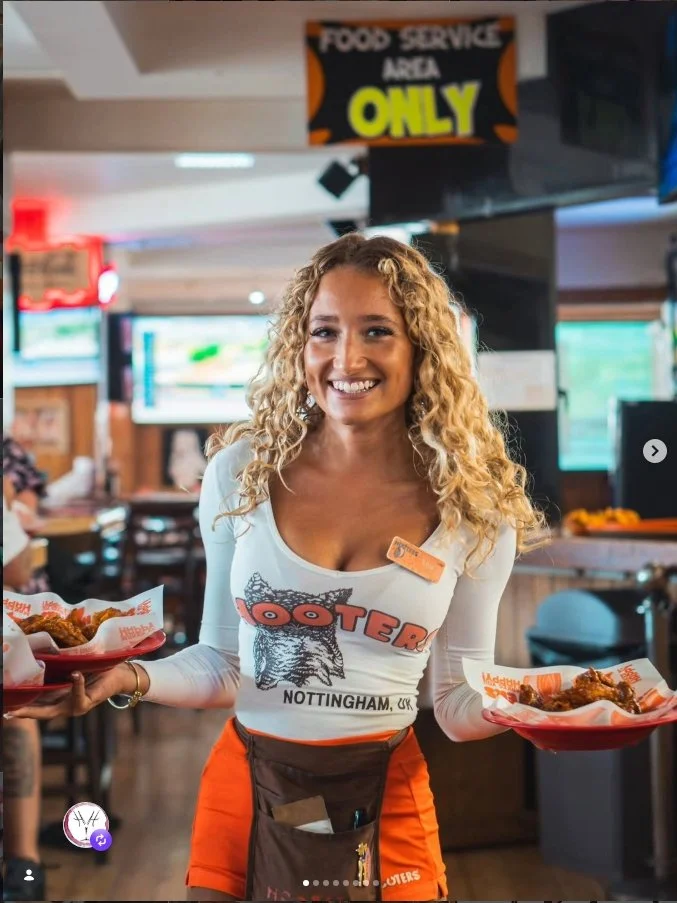 A smiling female employee at Hooters holding two trays of chicken wings at a restaurant. She is wearing a Hooters uniform with a name tag, a white long-sleeve shirt with the Hooters logo, and orange shorts.