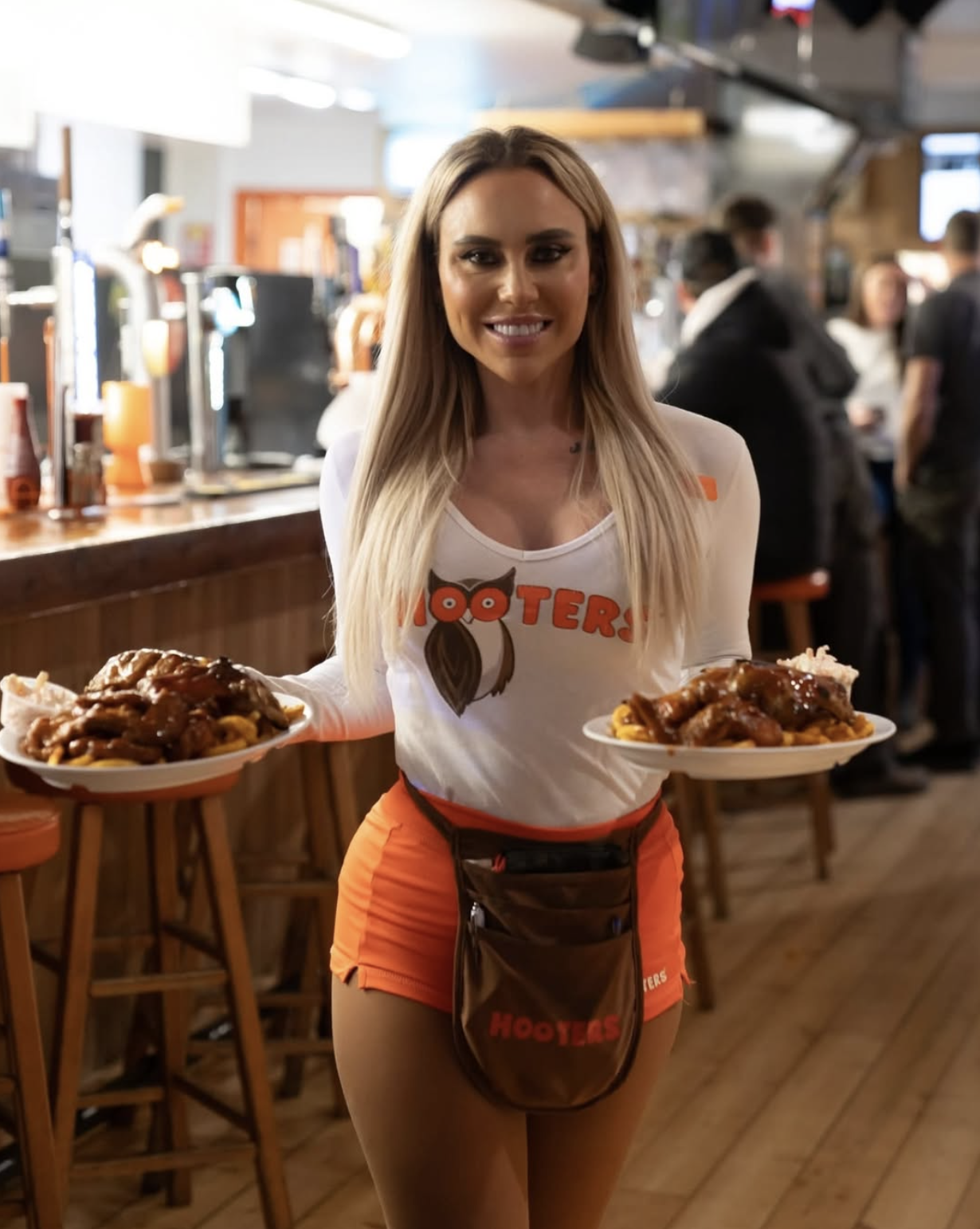A woman wearing a Hooters uniform holding two plates of food inside a restaurant. The uniform includes a white shirt with the Hooters logo, orange shorts, and a brown apron.
