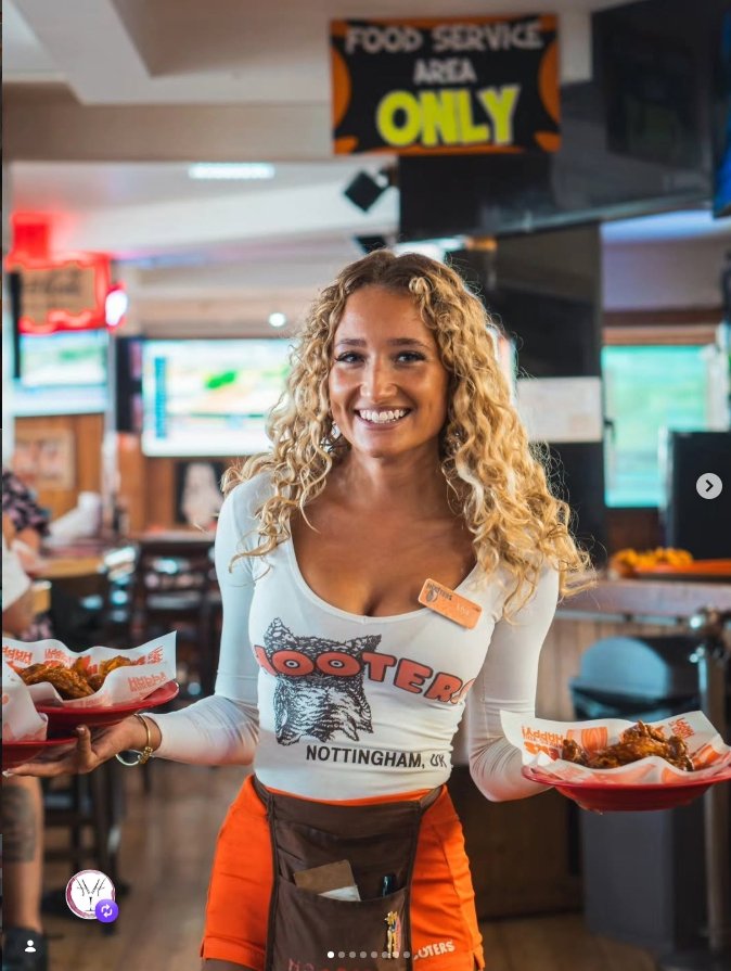 A woman smiling while holding two plates of food inside a restaurant, wearing a Hooters uniform with orange shorts and a white shirt with a logo.