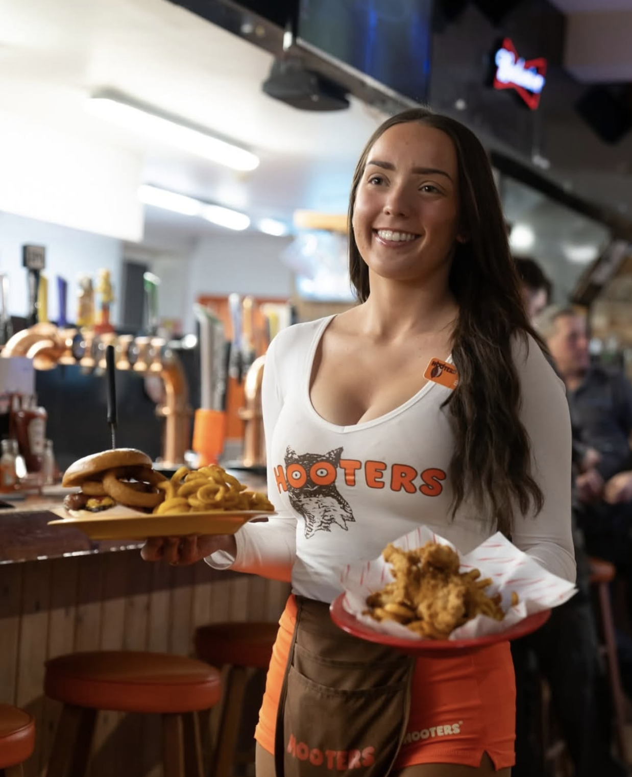 Hooters waitress smiling and holding plates of food in a restaurant