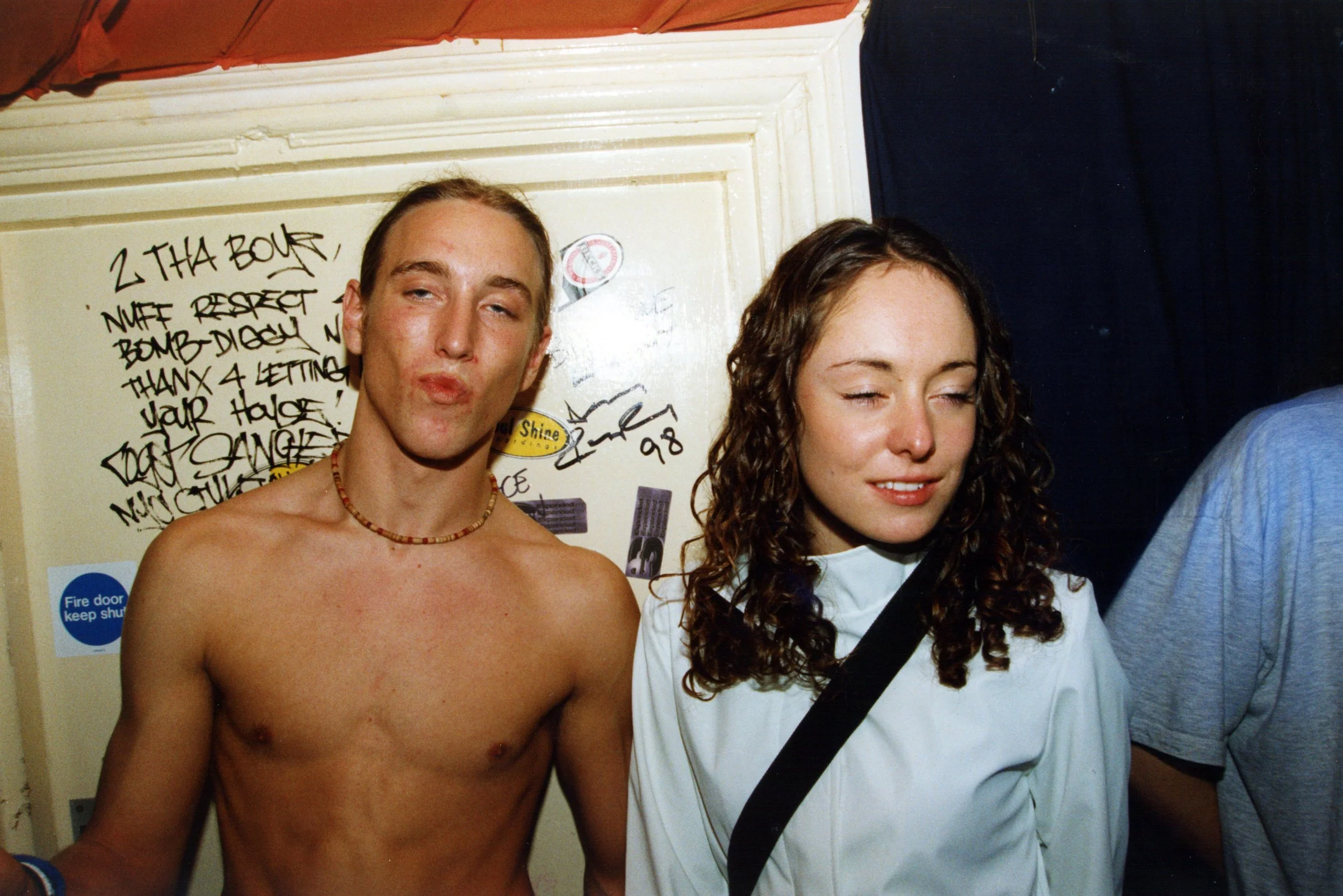 A shirtless young man with light skin and long hair, making a puckered kiss face next to a young woman with curly hair and light skin, who is looking down and smiling slightly. They are standing in front of a wall with handwritten notes and stencils,