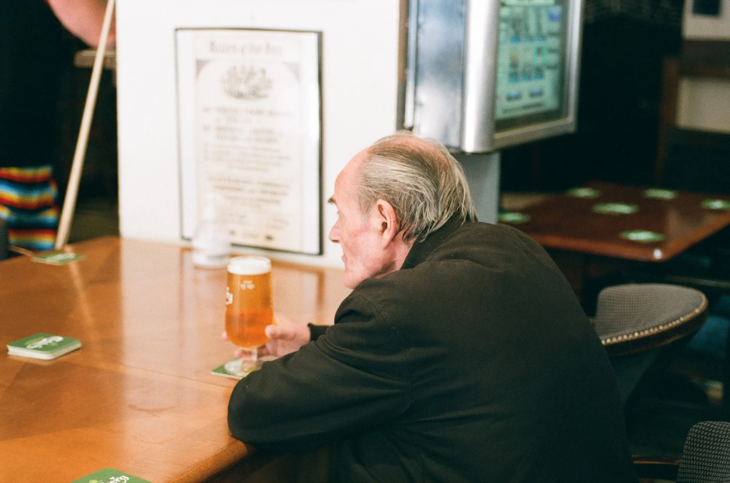 An older man with gray hair sitting at a wooden table, holding a glass of beer. There are some cards on the table, and in the background, a person with rainbow-colored clothing and a vending machine.