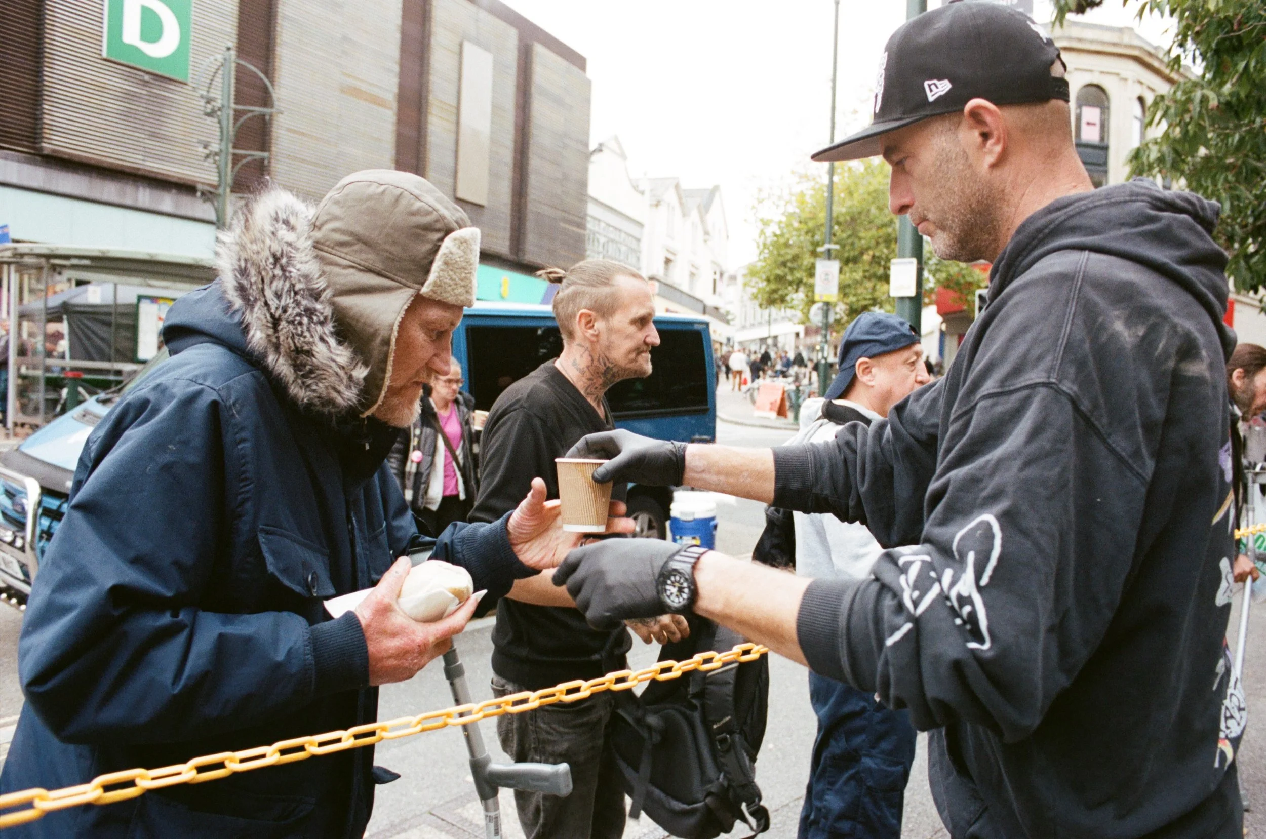 A homeless man with a beanie and coat receives a cup of coffee from a man wearing a black hoodie and cap on the street. Other people are visible in the background.