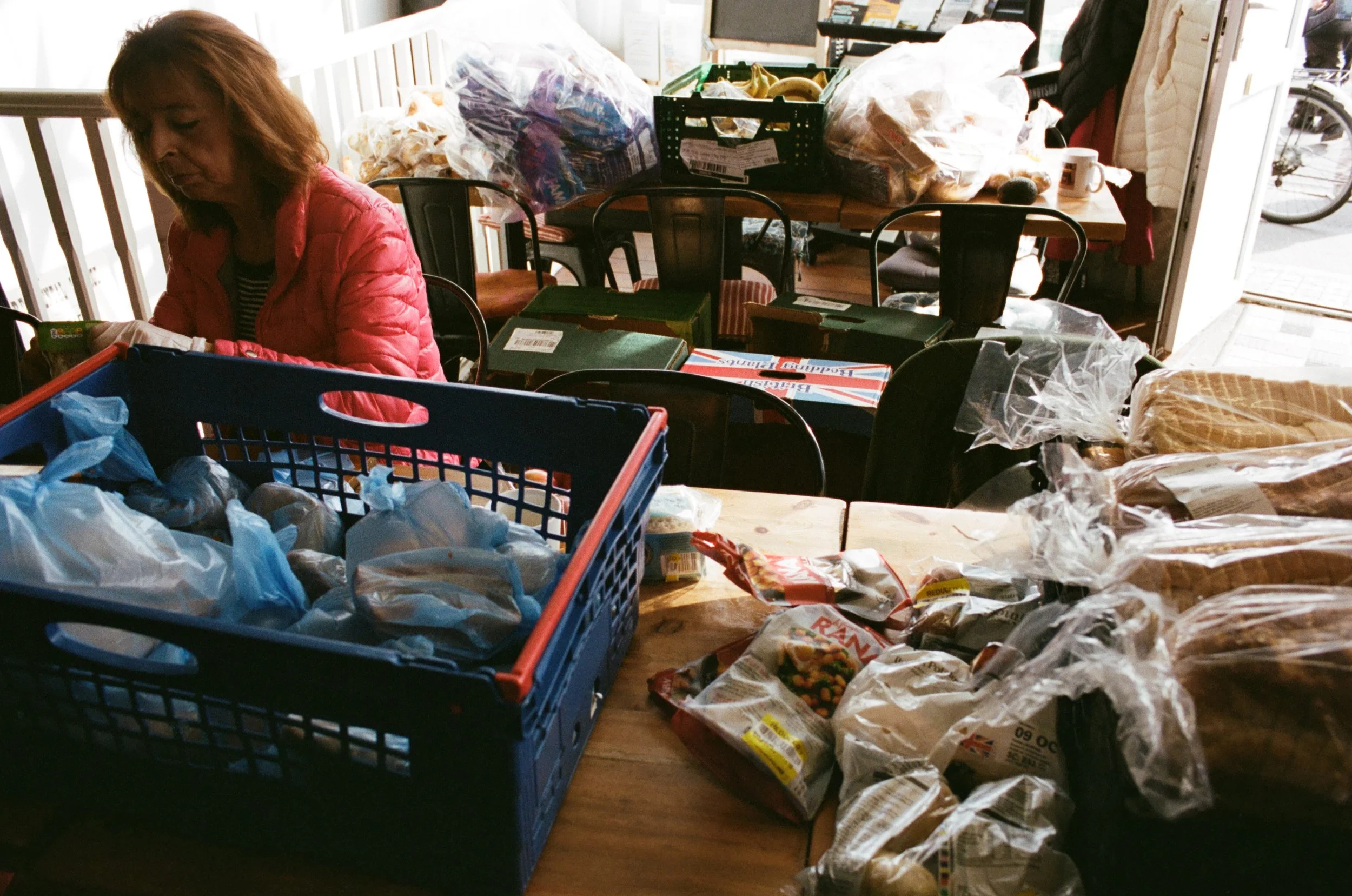 A woman with shoulder-length hair wearing a pink jacket is sitting at a cluttered dining table. The table is filled with various grocery bags, boxes, and packaged food. Behind her, there are more grocery items, a black crate, and a partially open doo