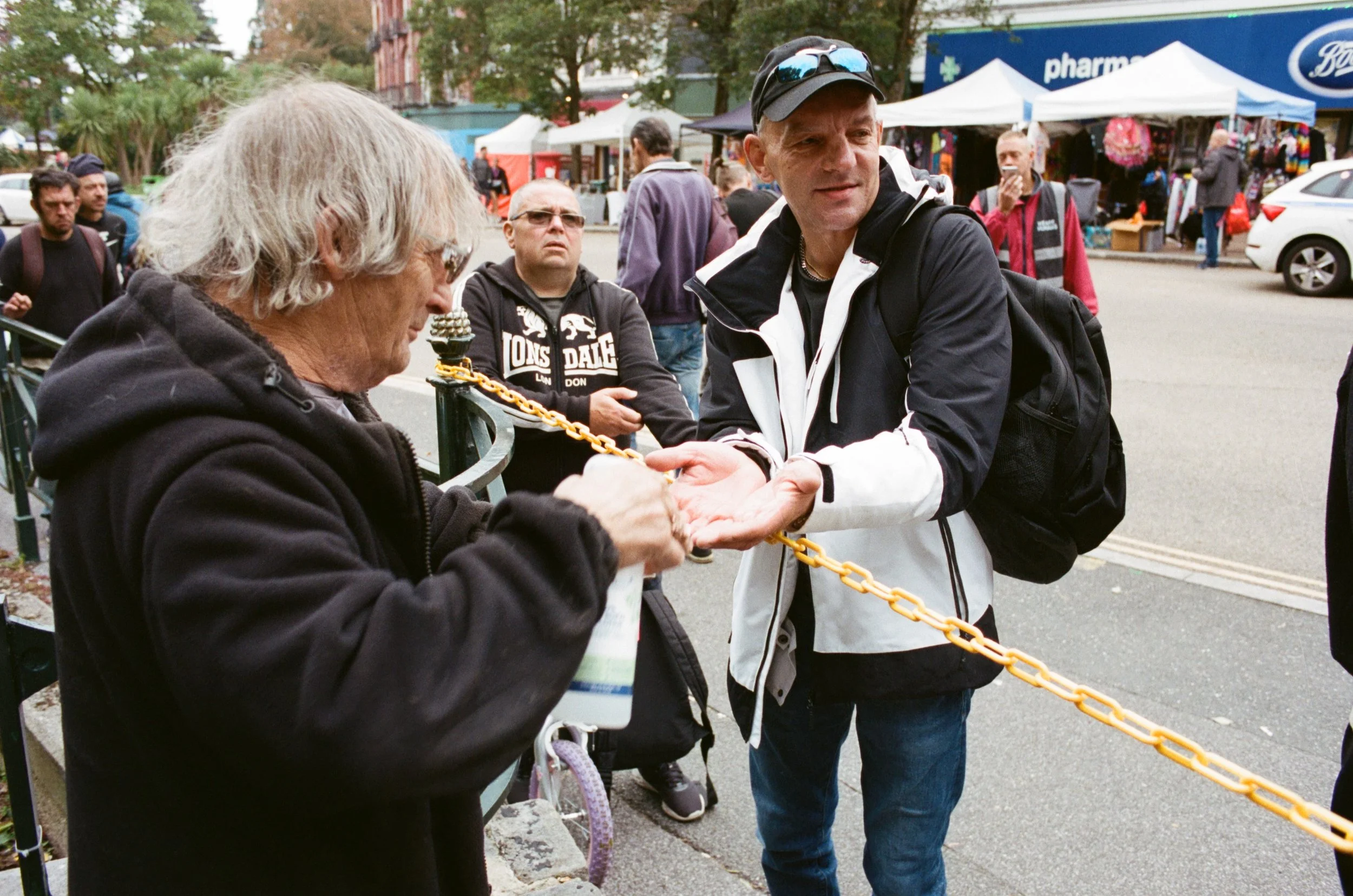 A man shakes hands with an elderly woman at an outdoor market, with other people and market stalls in the background.