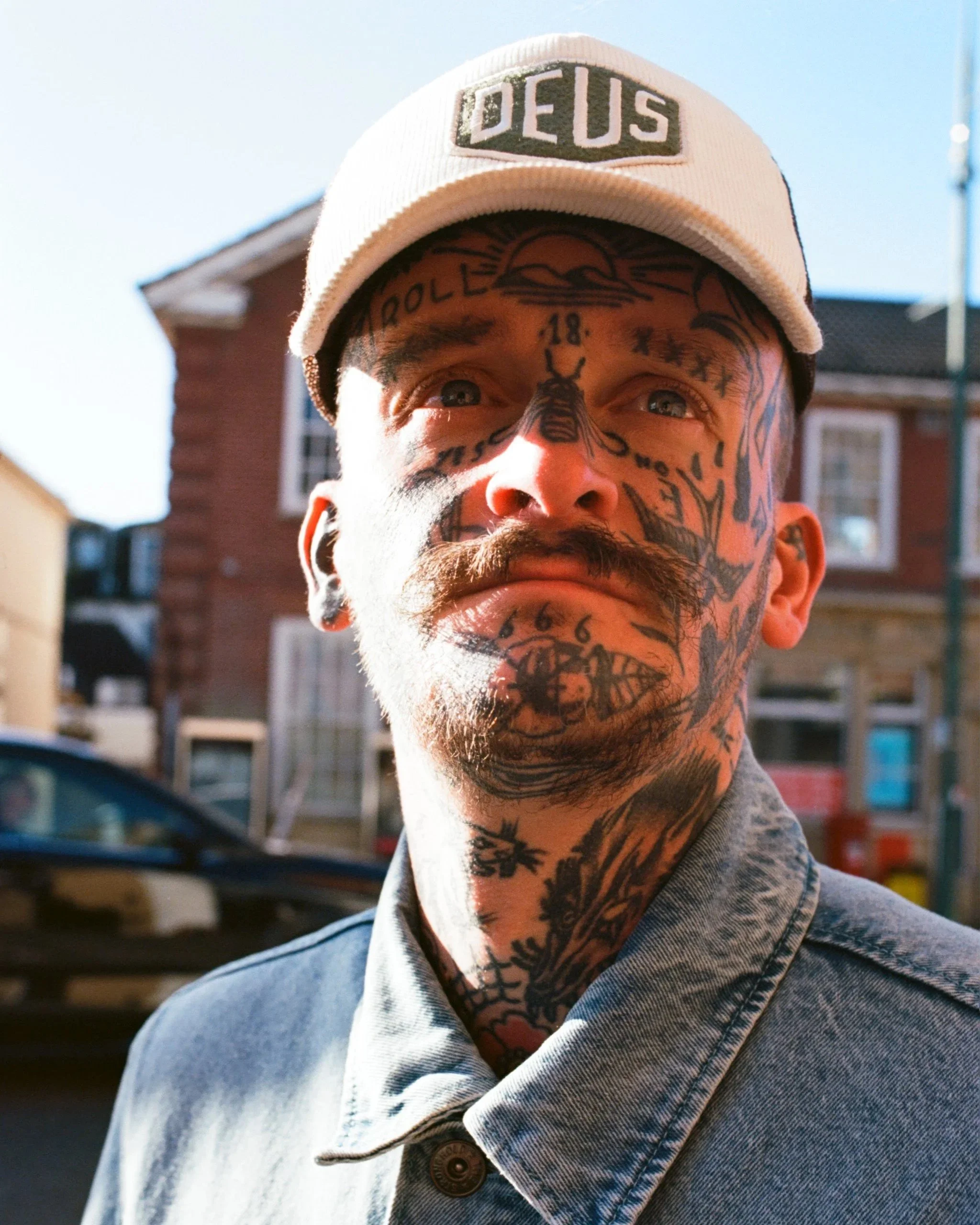 Close-up of a man with tattoos, wearing a beige cap with 'DEUS' embroidered, and a denim jacket, standing outdoors in front of residential buildings.