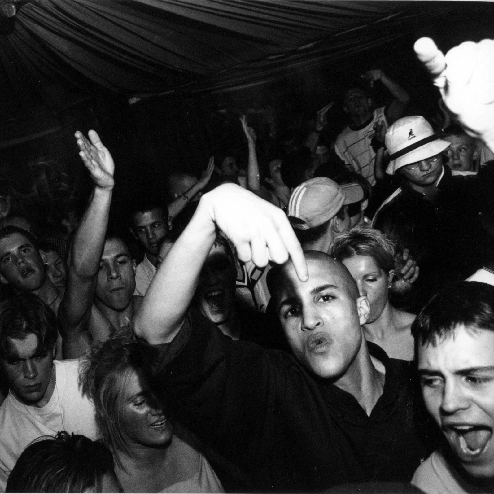 A black and white photo of a crowded party or club with young people dancing and socializing. Some individuals are raising their hands and making gestures, with a mix of facial expressions including smiling and talking.
