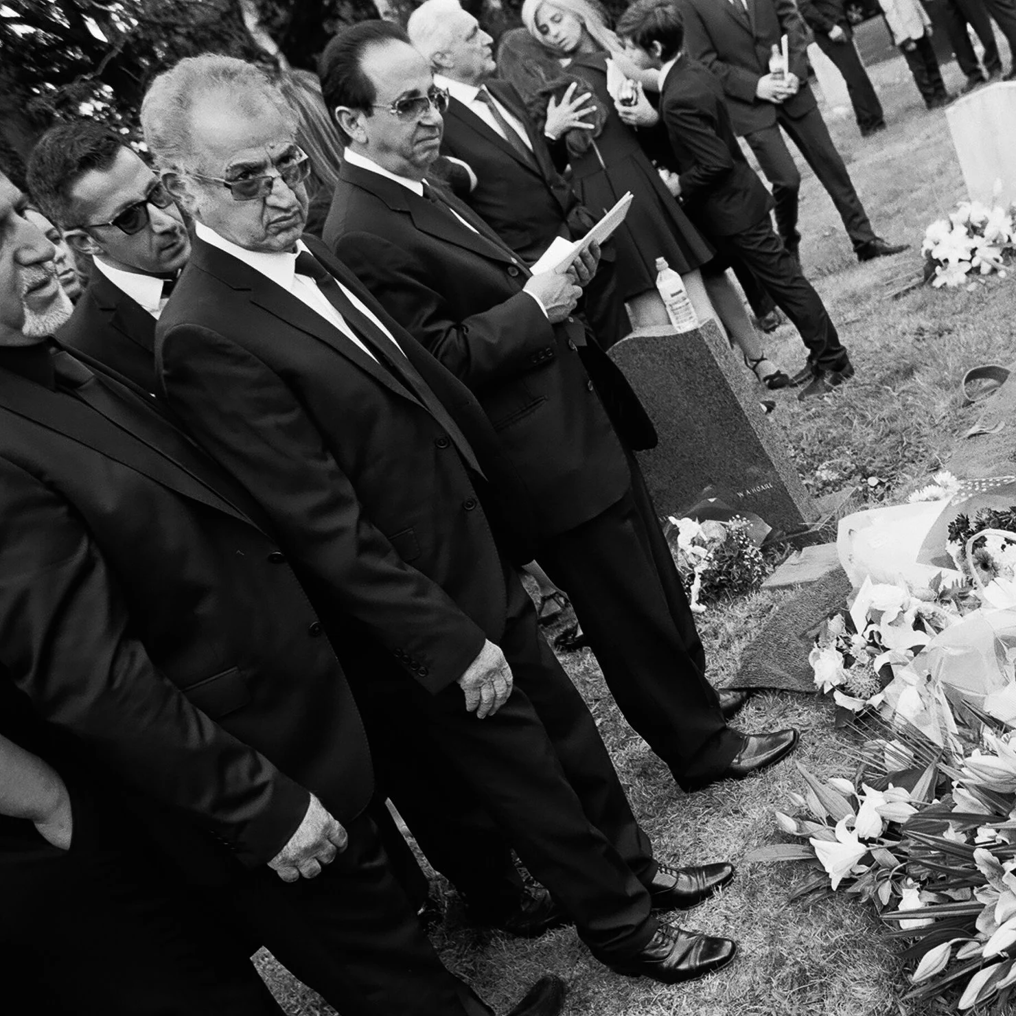 A group of men in suits and sunglasses standing solemnly during a funeral, paying respects at a gravesite with flowers and wreaths.