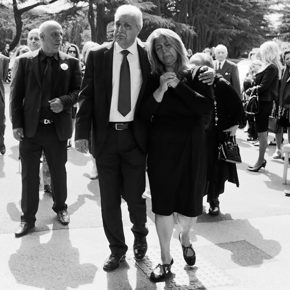 A black-and-white photo of a mourning scene with several people dressed in black, some with somber expressions. A woman appears to be grieving, leaning on a man as they walk through a funeral procession.