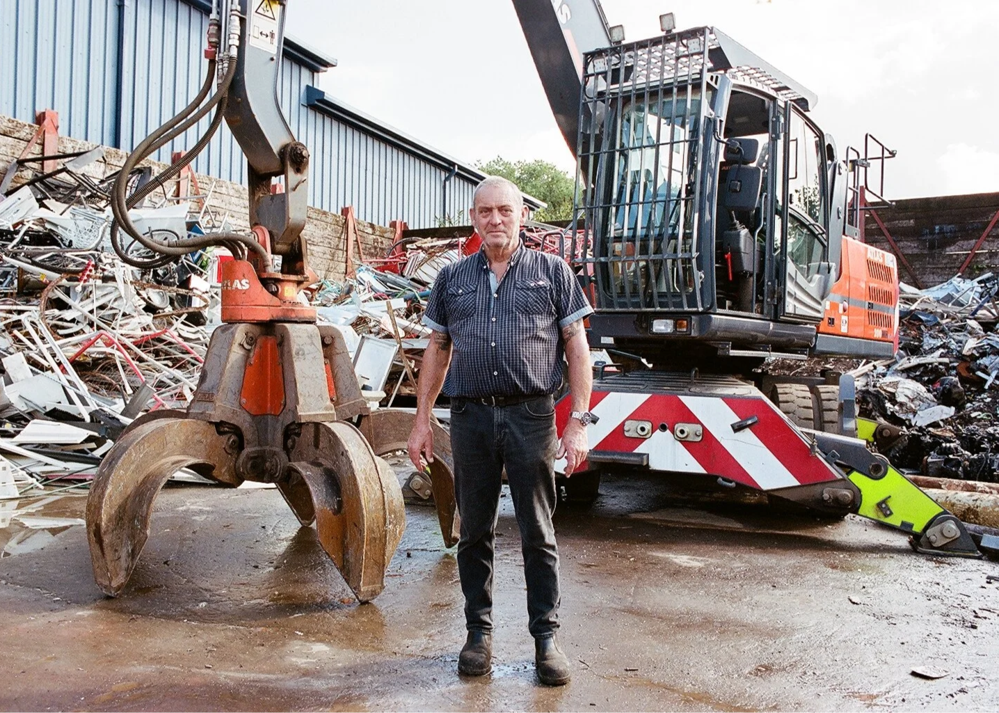 A man standing in front of scrap metal and construction equipment, including a large orange excavator and a metal grapple in a scrapyard.