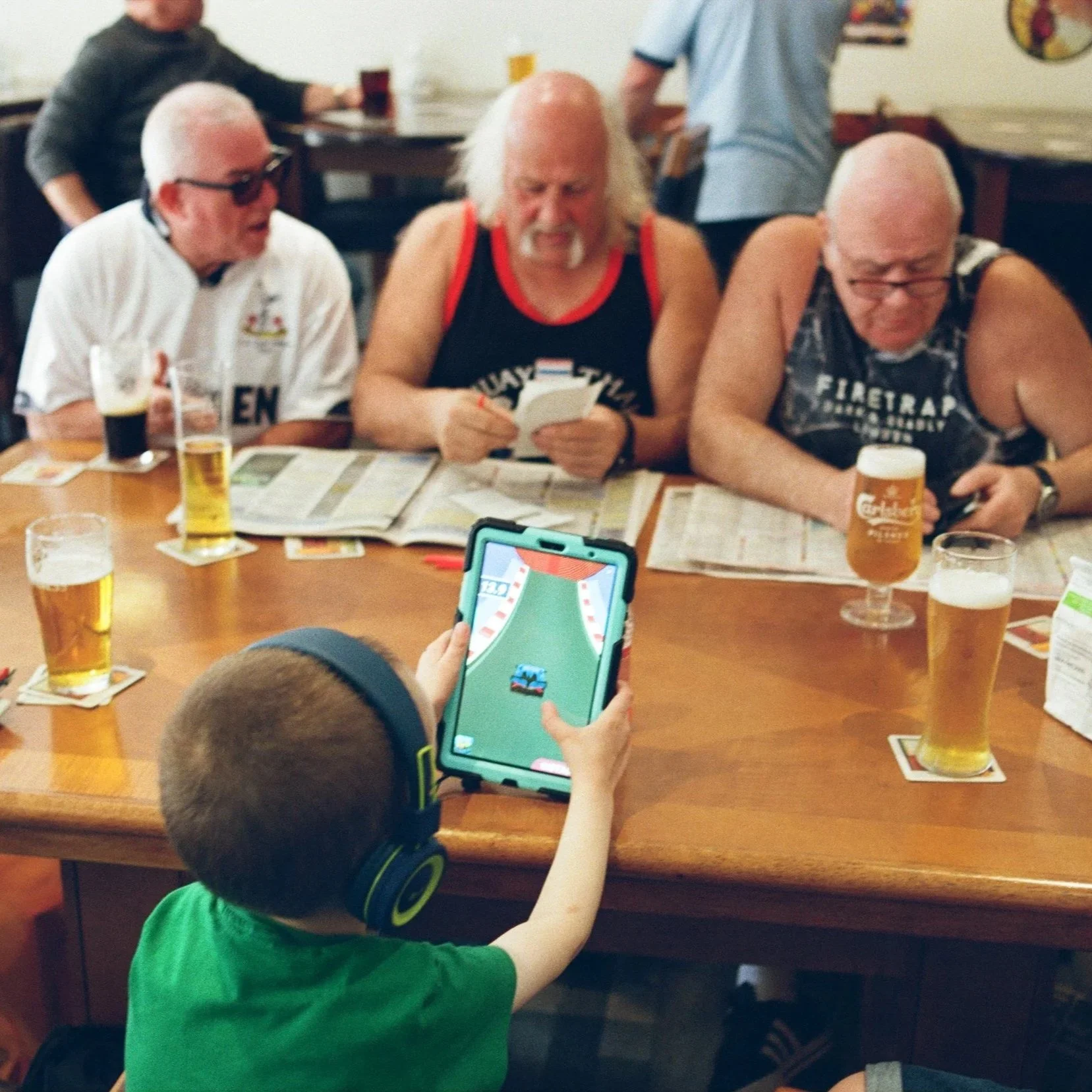 A group of three elderly men sitting at a wooden table in a restaurant, playing a puzzle game on a tablet, with beers in front of them and a boy in a green shirt and headphones in the foreground, playing a game on a tablet.