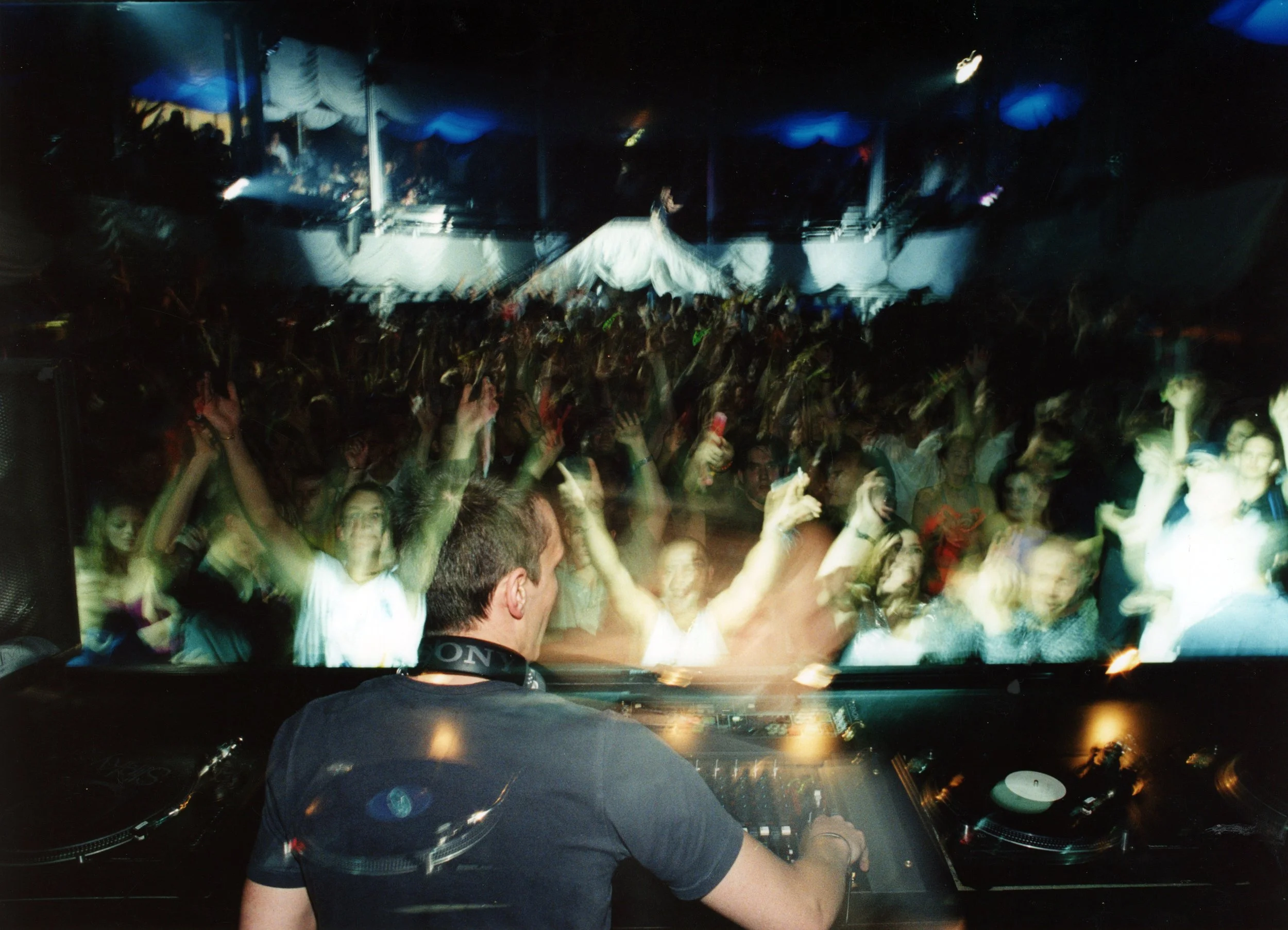 A person wearing a black shirt and headphones sitting at a DJ console, with a crowd of people dancing and raising their hands in a dimly lit club or concert setting.