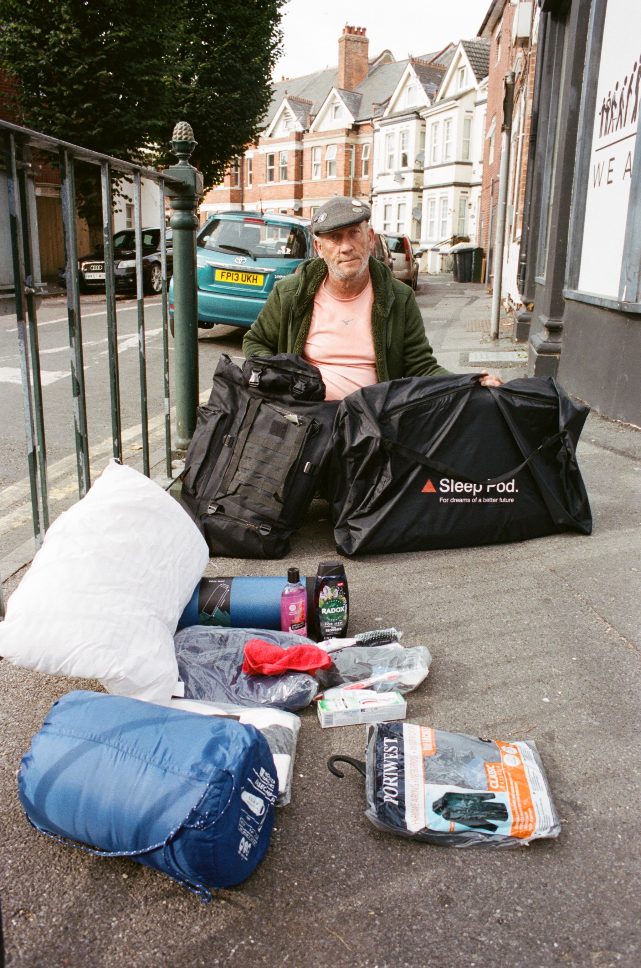 A man sitting on the sidewalk behind a metal fence with various camping and outdoor gear spread out in front of him, including a sleeping bag, backpack, and bottles, with residential houses and parked cars in the background.