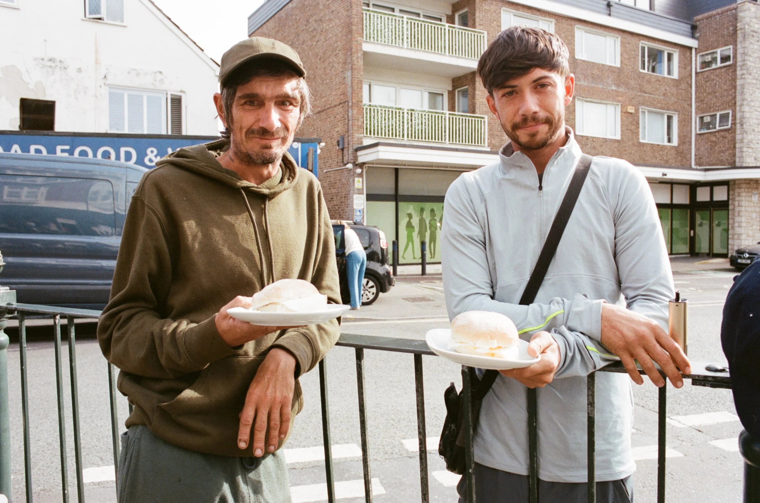 Two men standing outside on a city street, each holding a plate with food, with buildings and parked cars in the background.