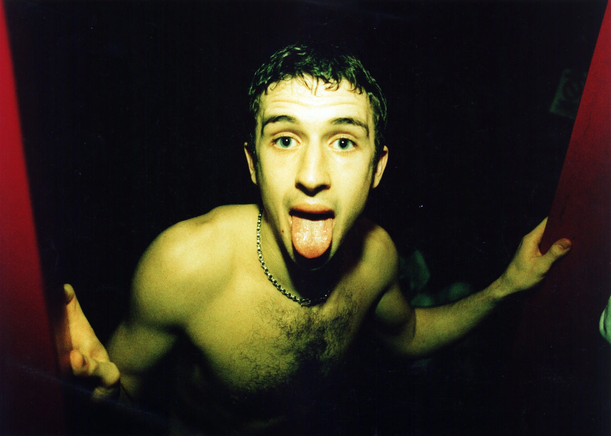 A topless young man with short dark hair and blue eyes sticking out his tongue and looking directly at the camera. He is wearing a silver necklace and appears to be taking a selfie in a dark room with a red and black background.