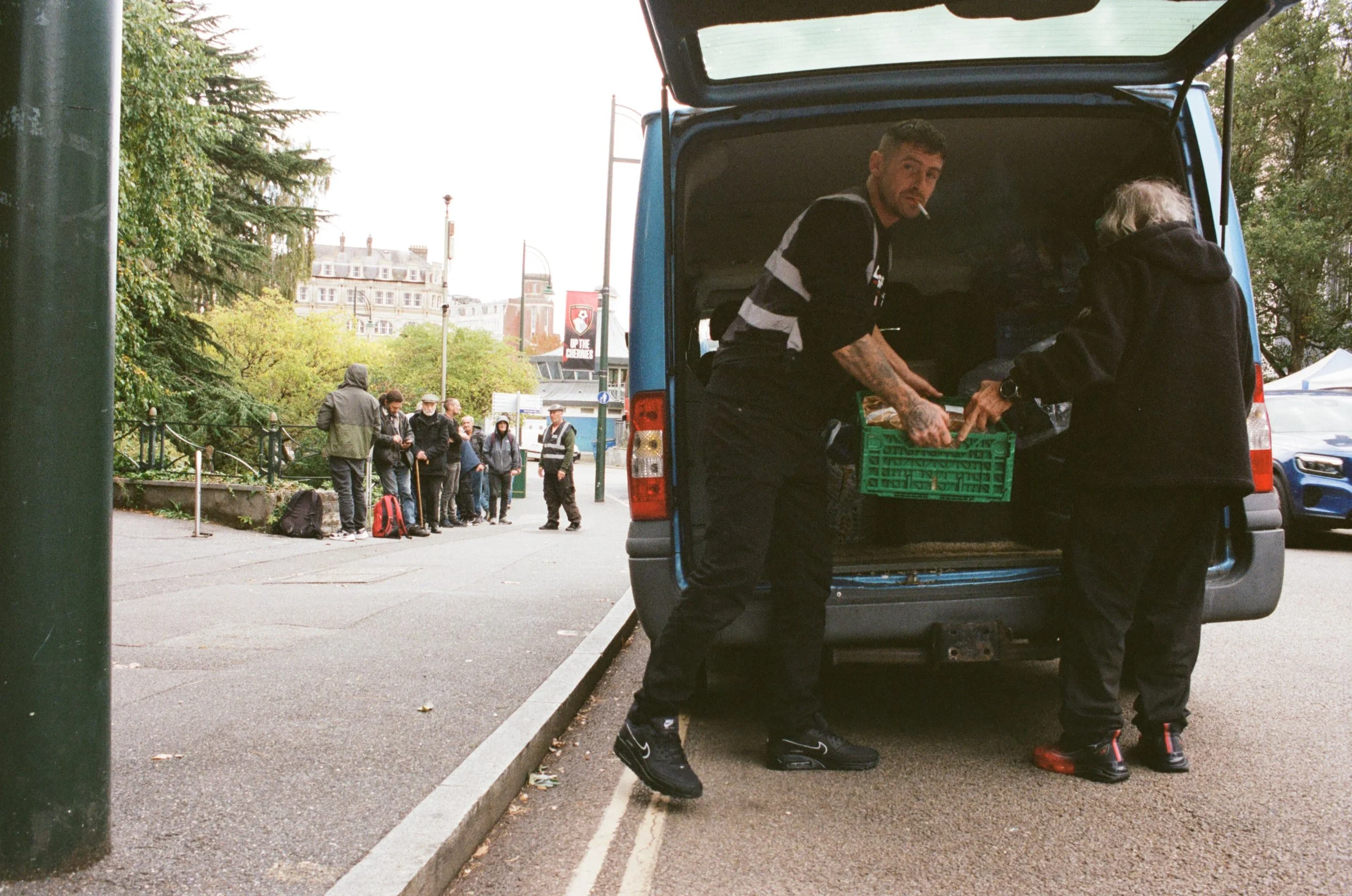 Man is unloading groceries from a blue van, which has its back doors open, while a woman stands nearby. Several people are visible in the background waiting in line on a city street with trees and buildings.