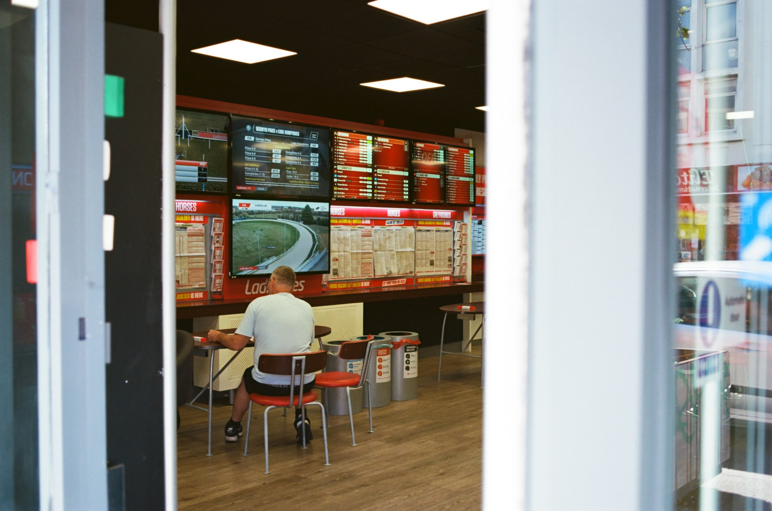 View through a glass door shows a betting office with multiple electronic screens displaying horse racing information. Inside, a man sits at a table, and there are chairs, posters, and other typical betting shop fixtures.
