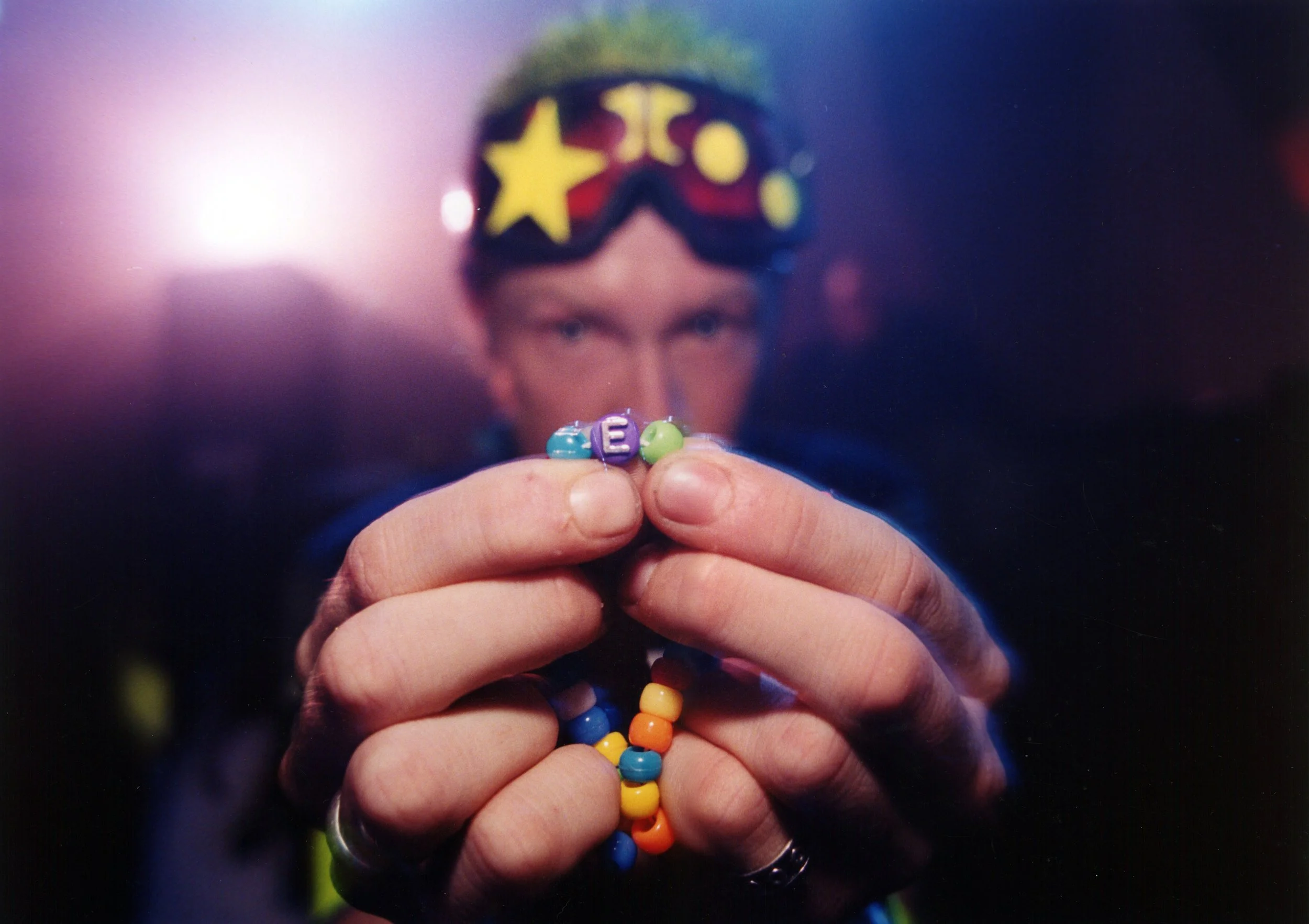 Close-up of a person holding colorful beads spelling out the letter 'E' against a blurred background with a person wearing sunglasses, which have yellow star and circle decorations on the lenses.